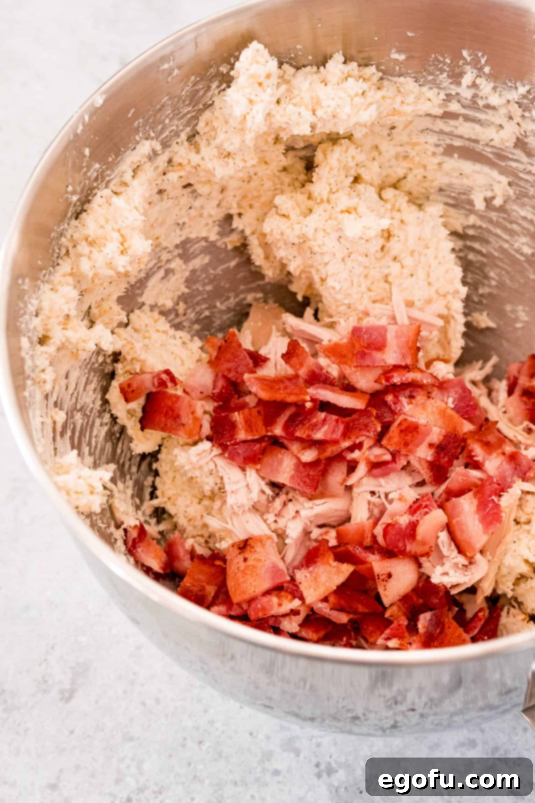 Crispy chopped bacon pieces added to the creamy cheese and ranch seasoning mixture in a mixing bowl, ready to be incorporated for the Crack Chicken Ball filling.