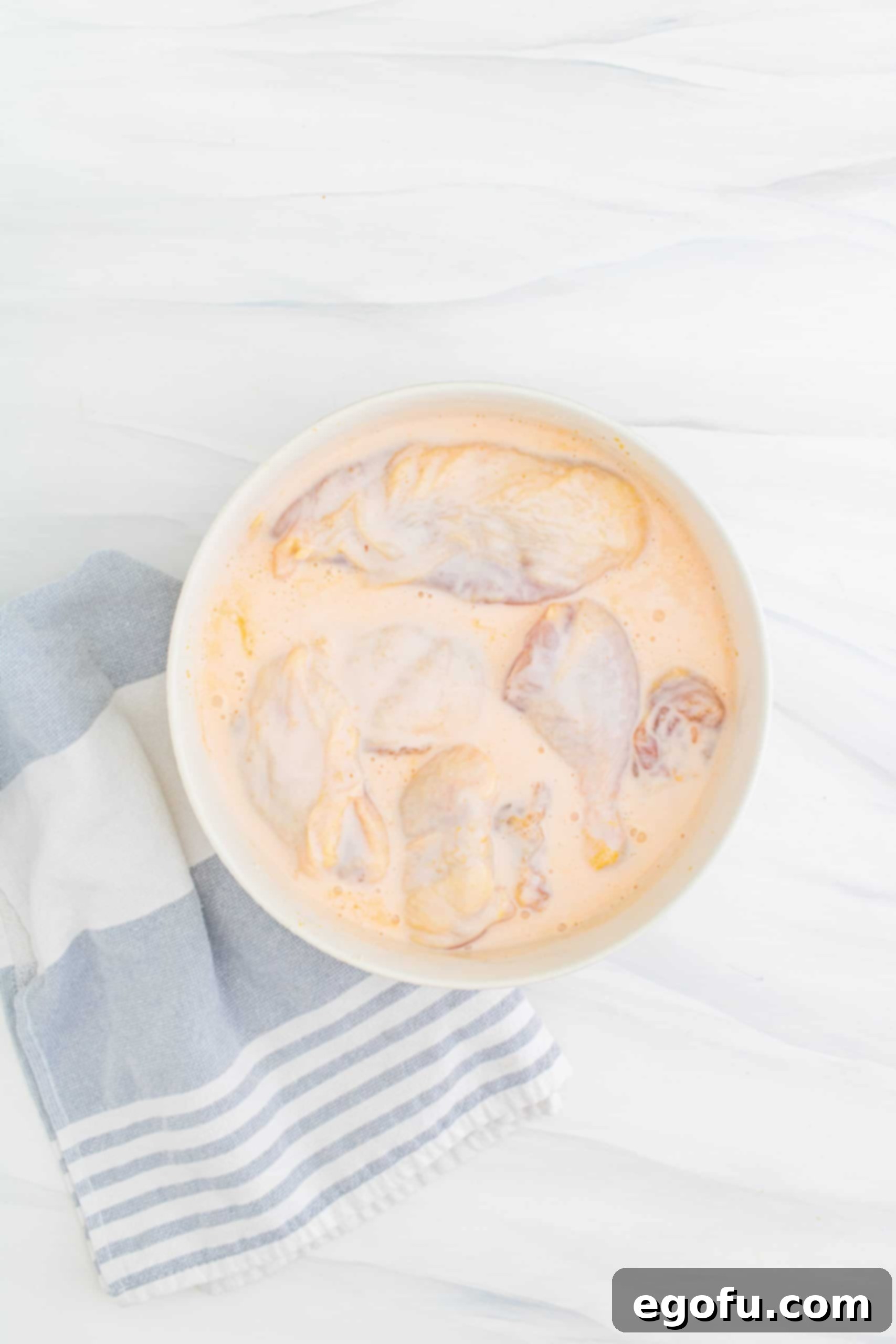 chicken pieces soaking in hot sauce and buttermilk in a white bowl.