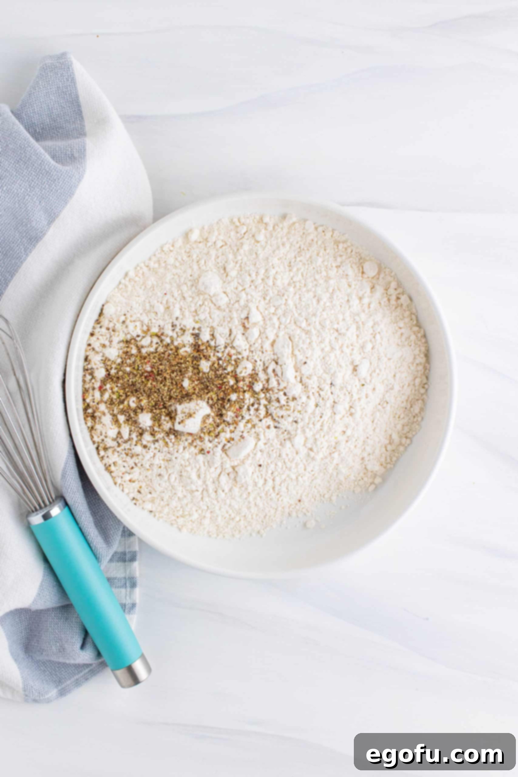 Flour, cornstarch and black pepper in a white bowl with a whisk set off to the side.