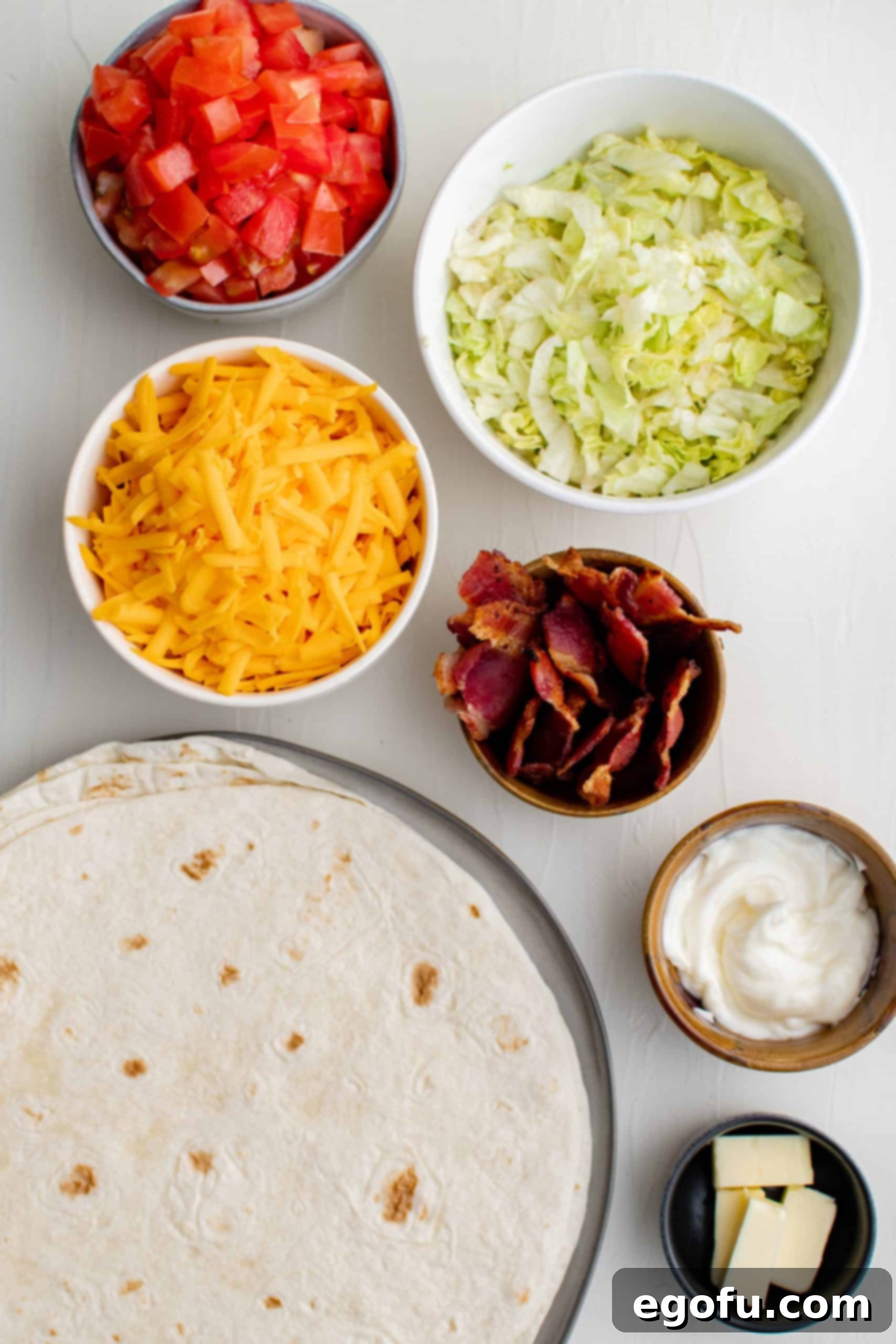 Diced tomatoes, shredded cheddar cheese, shredded lettuce, cooked bacon, and flour tortillas laid out as BLT Quesadilla ingredients.