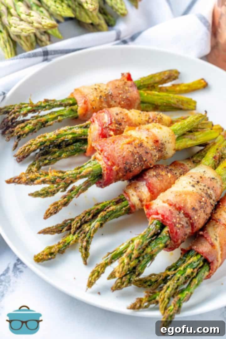 Crispy Air Fryer Bacon Asparagus Bundles 10 Six bacon wrapped asparagus bundles laying on a round white plate with fresh asparagus in the background.