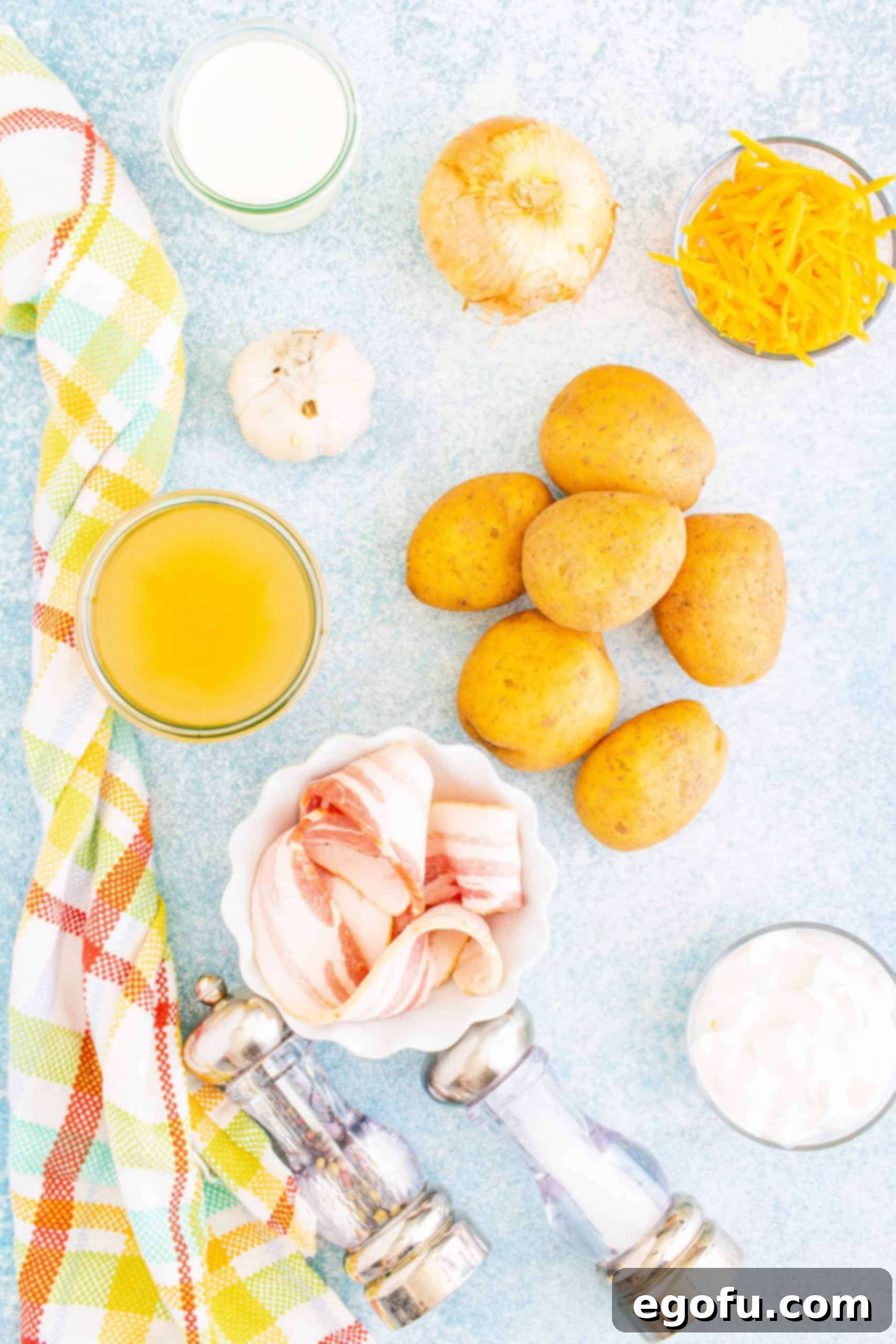 Arrangement of key Instant Pot Loaded Potato Soup ingredients: yellow potatoes, uncooked bacon, garlic cloves, block of cheddar cheese, chicken stock carton, diced onion, heavy cream, salt, and pepper.