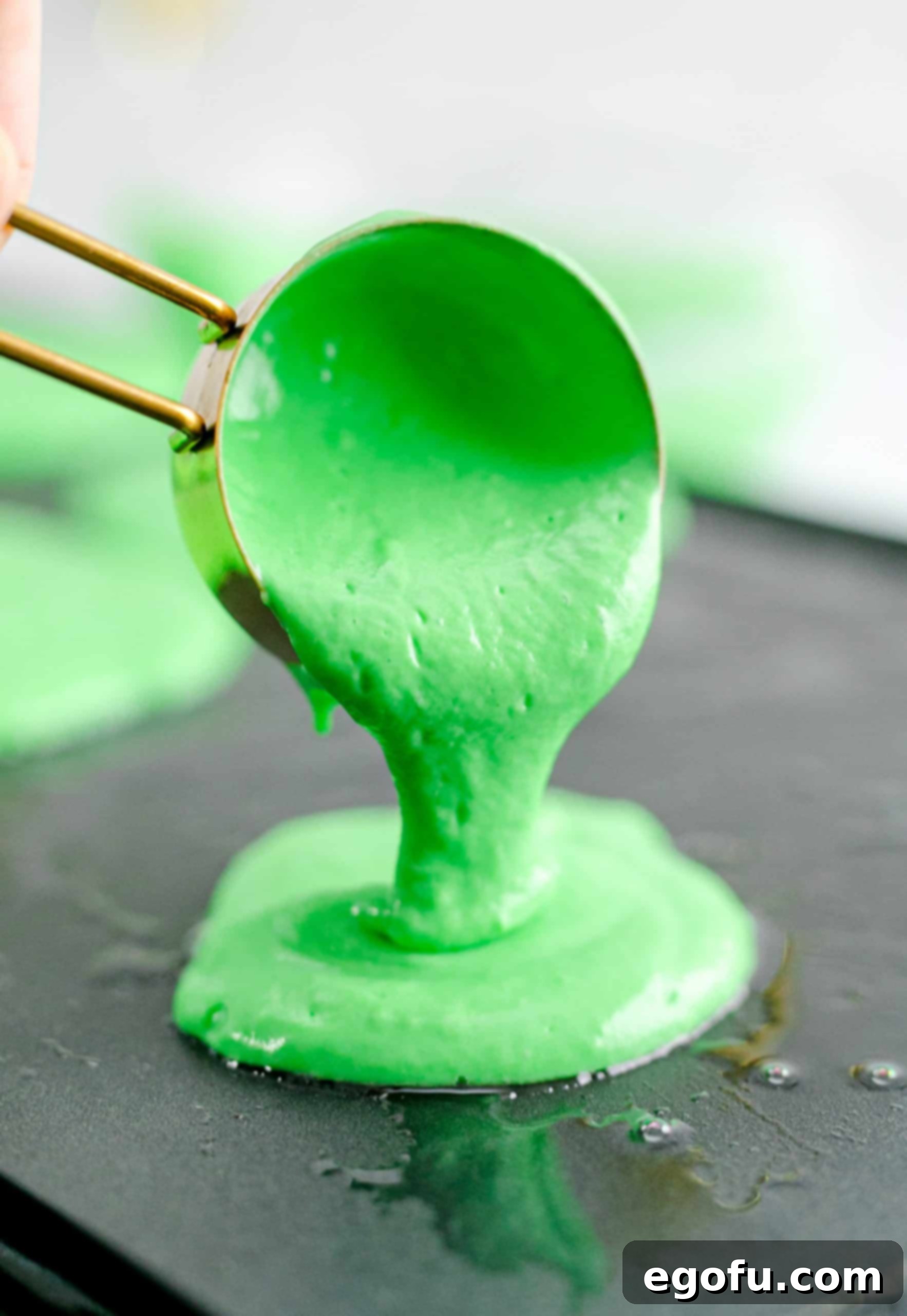 Green pancake batter being poured from a measuring cup onto a hot griddle, starting the cooking process for a St. Patrick's Day breakfast.