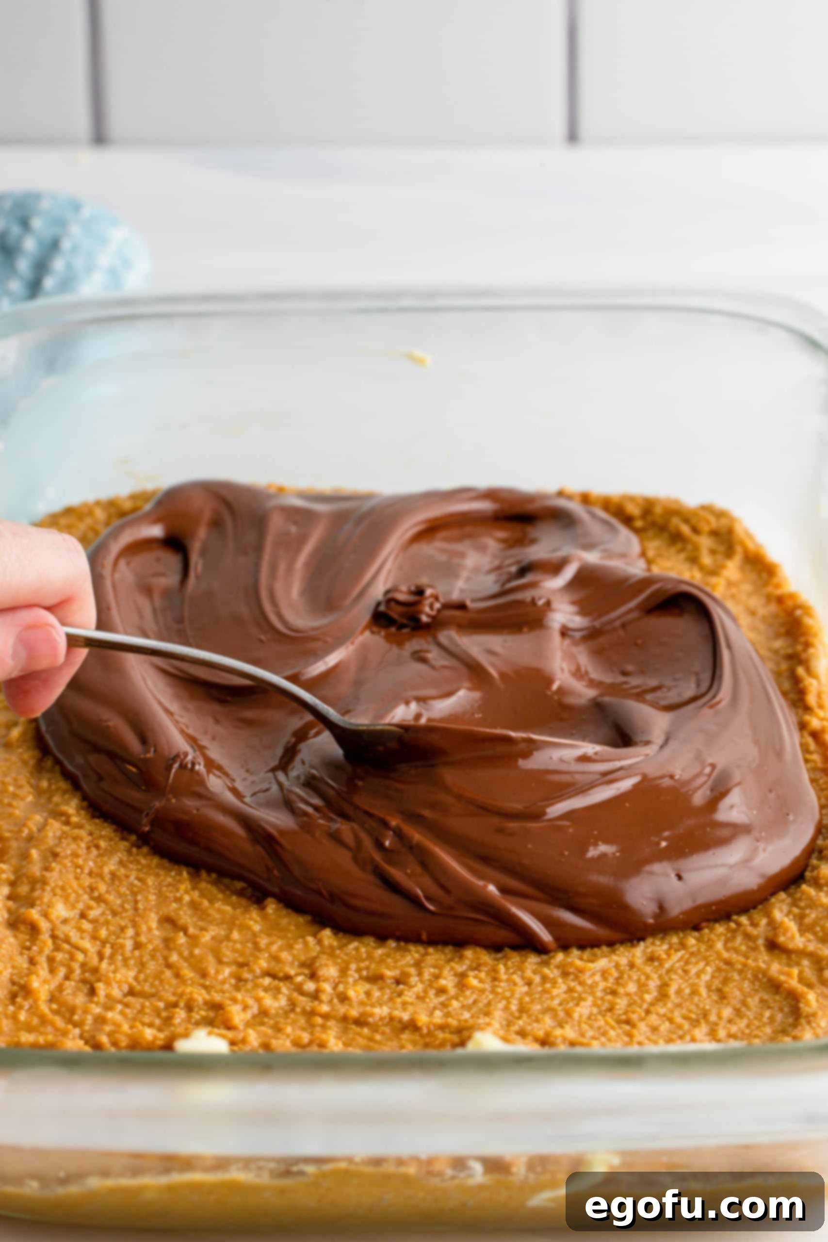 Grandma's No-Bake Peanut Butter Dream Bars 11 A spoon spreading the warm, melted chocolate evenly over the chilled peanut butter layer in the baking dish.