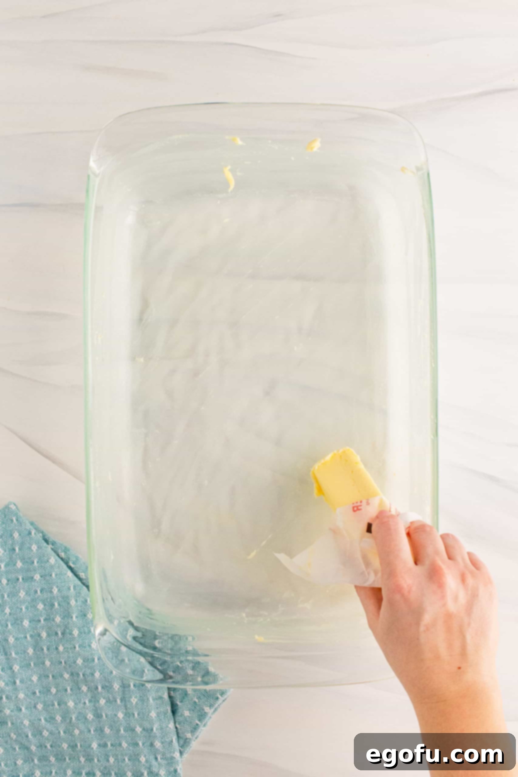Grandma's No-Bake Peanut Butter Dream Bars 5 A hand buttering a glass baking dish with a stick of butter, preparing it for the peanut butter bar mixture.