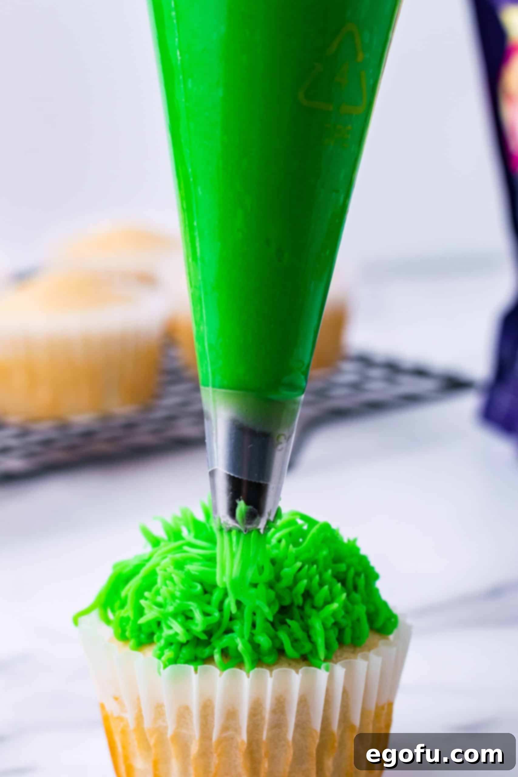 A baker expertly piping green frosting onto the top of a cupcake to create a grass-like effect.