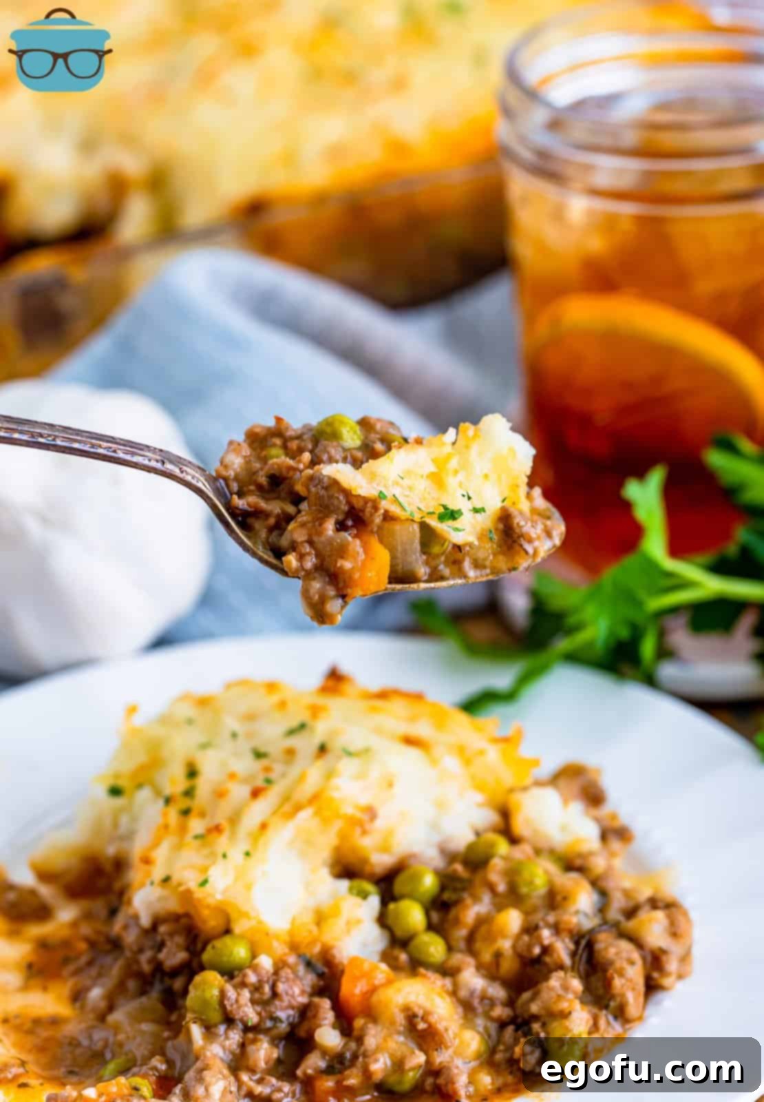 A delectable serving of Shepherd's Pie artfully presented on a white plate, with a fork lifting a portion to showcase its creamy topping and rich filling, complemented by a refreshing glass of iced tea in the soft-focus background.