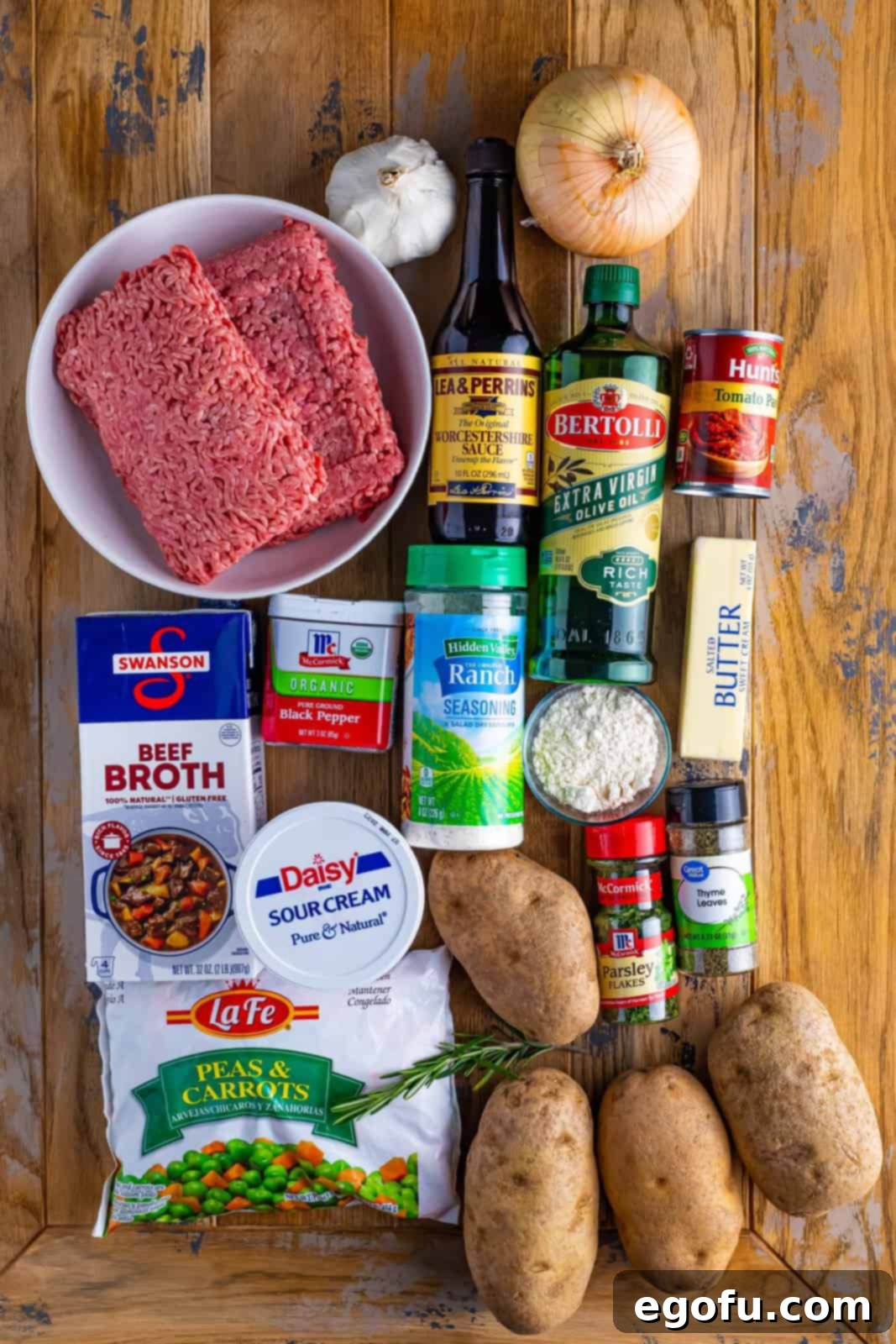 A vibrant flat lay of all the fresh and pantry ingredients needed for Shepherd's Pie, including olive oil, onion, parsley, rosemary, thyme, pepper, ground beef or lamb, garlic, Worcestershire sauce, flour, tomato paste, frozen peas and carrots, beef broth, potatoes, sour cream, butter, and ranch seasoning.
