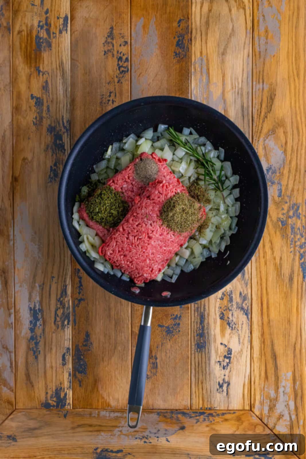 Ground beef browning in a skillet with sautéed onions, infused with dried thyme, fragrant rosemary, and cracked black pepper, creating a rich and aromatic base for the pie.
