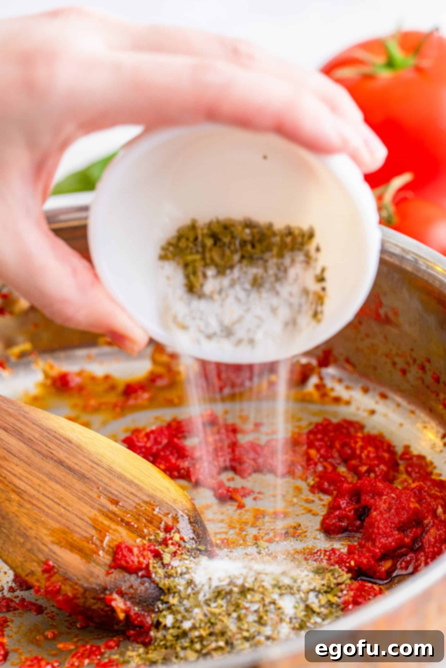 From Scratch Pizza Sauce Perfection 7 Granulated sugar, dried oregano, kosher salt, and black pepper being added to the simmering tomato and garlic base in the pan, ready for thorough mixing.