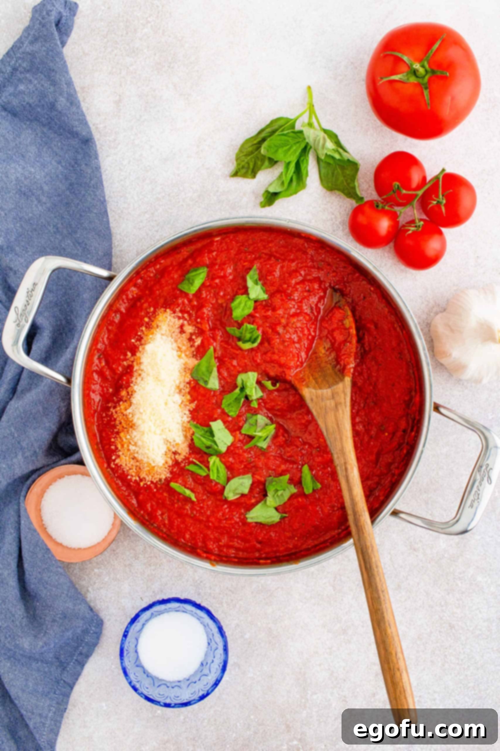 From Scratch Pizza Sauce Perfection 8 Overhead view of homemade pizza sauce in a pan, freshly removed from heat, with vibrant torn basil leaves and a sprinkle of grated parmesan cheese waiting to be stirred in.