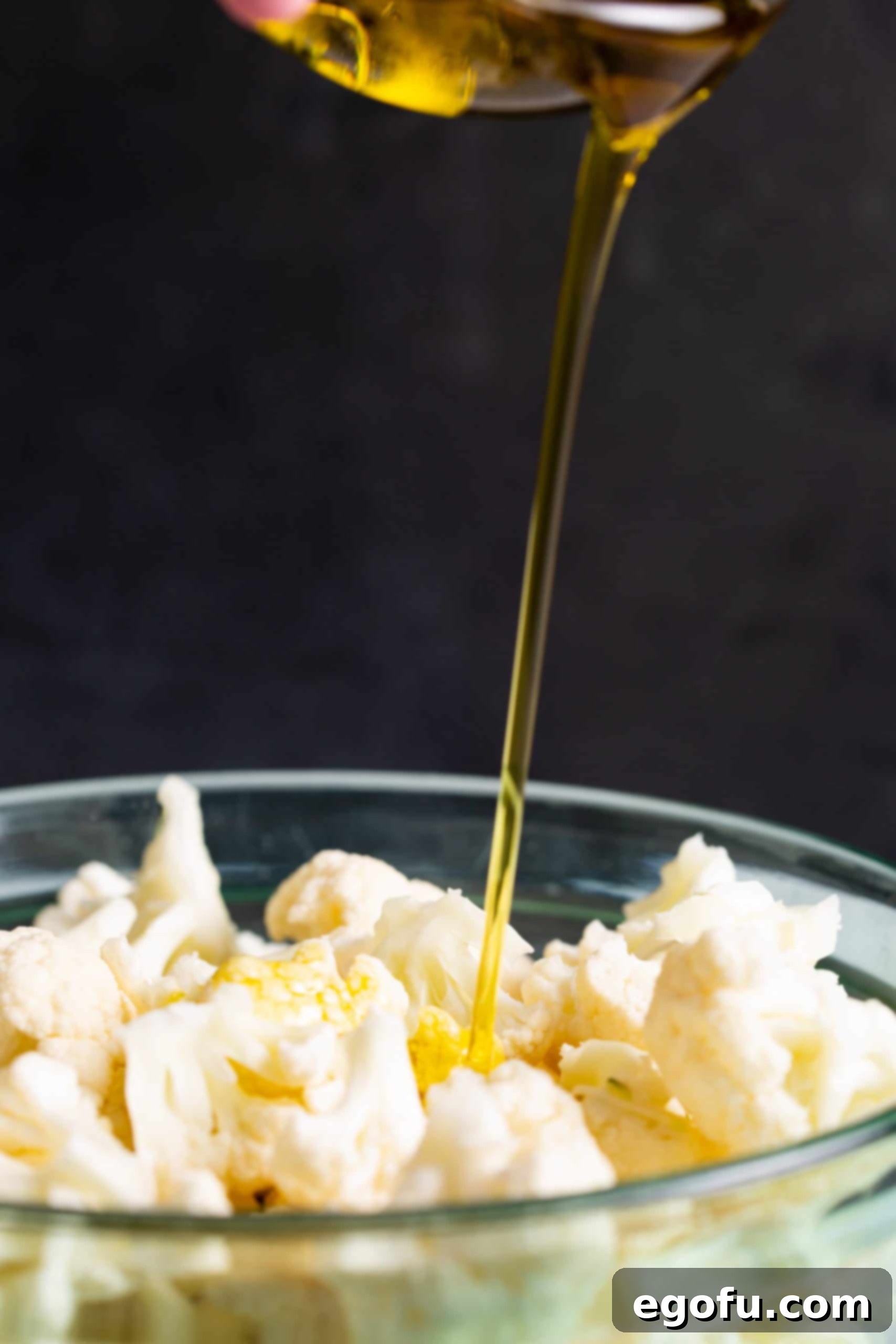 Olive oil being drizzled over cauliflower florets in a large mixing bowl, ready for seasoning.
