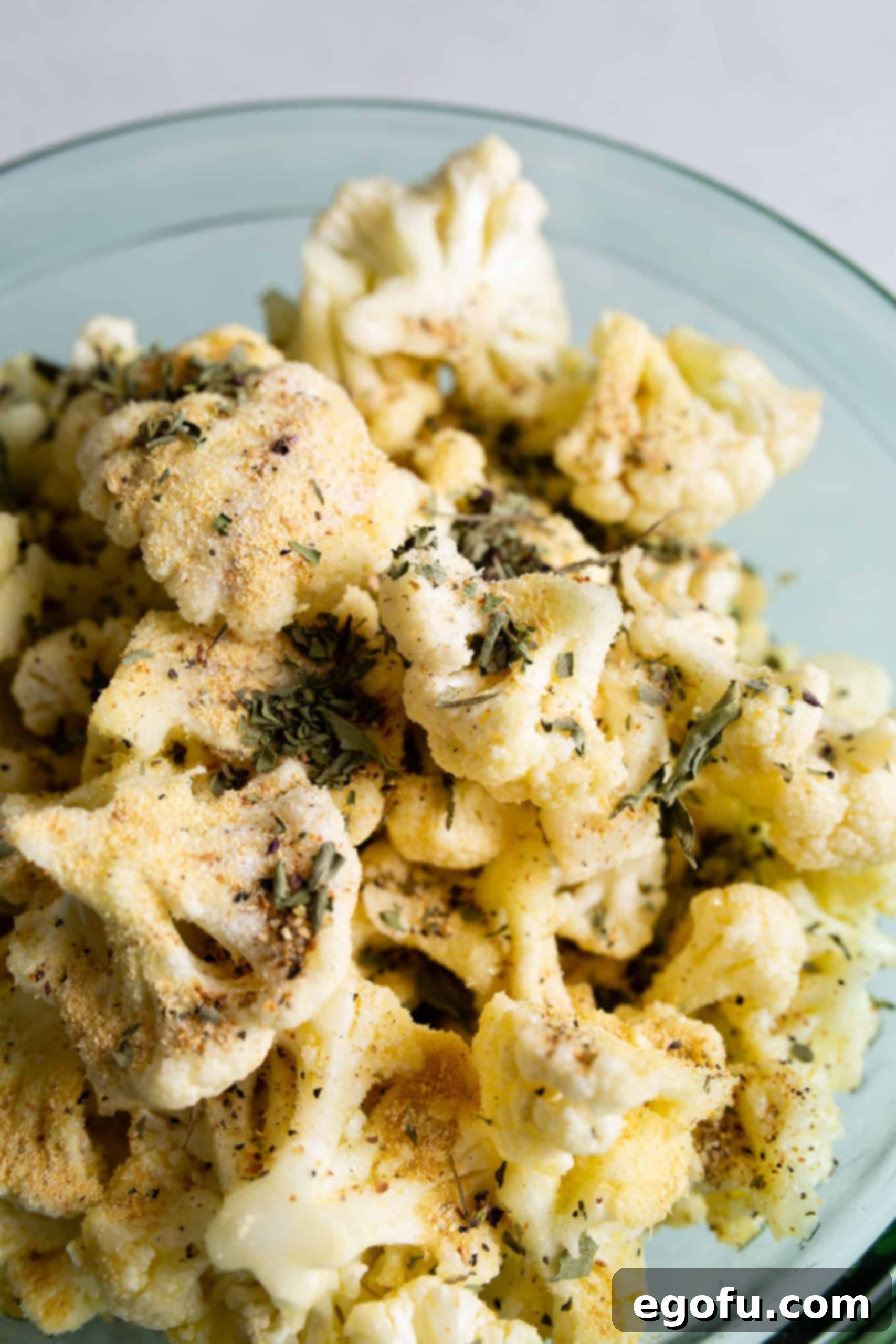 Dry spices and herbs being added to the oil-coated cauliflower florets in a bowl, ready for mixing.