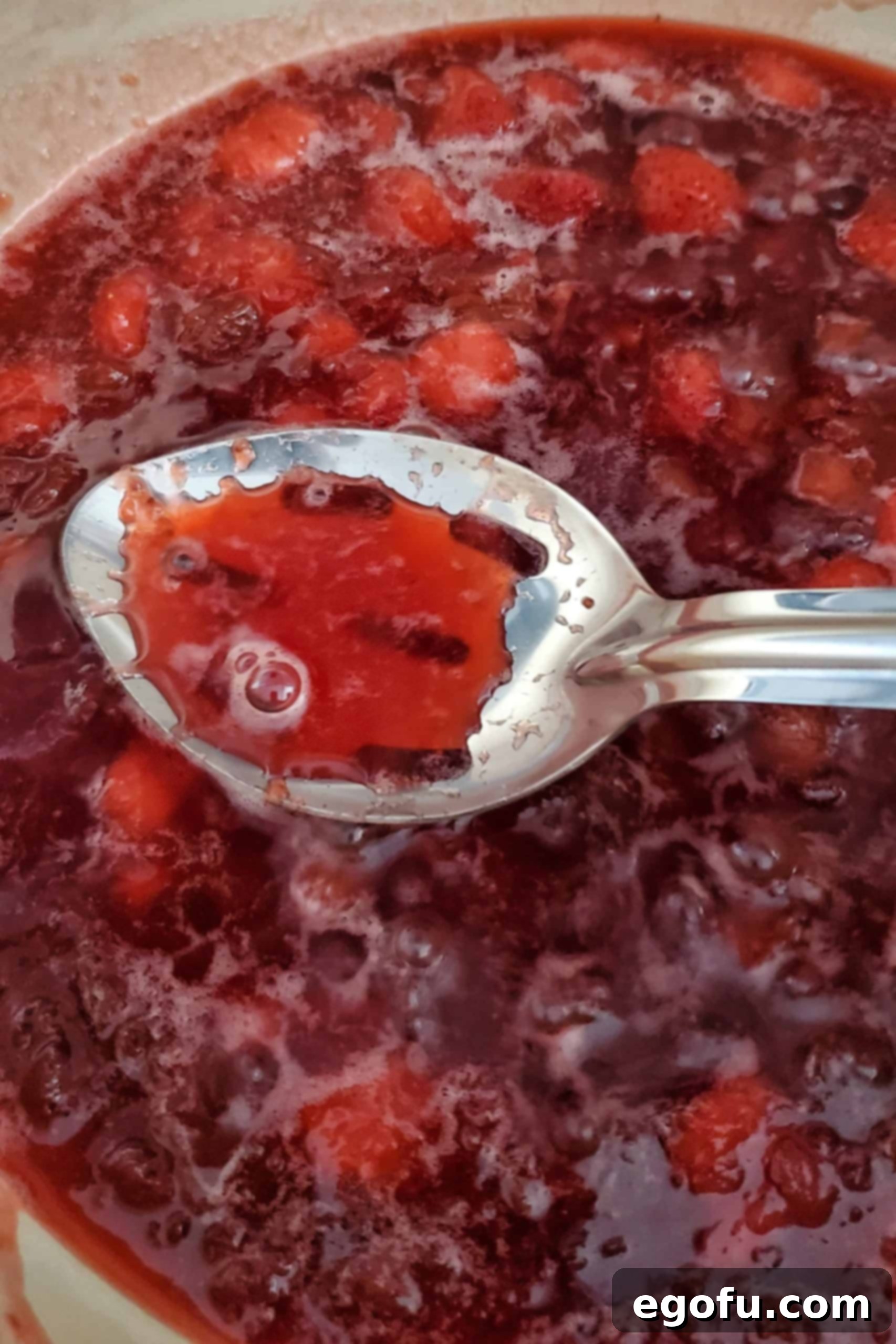 A spoon lifting a sample of the simmering jam from the saucepan, showing its reduced liquid and chunky fruit consistency.