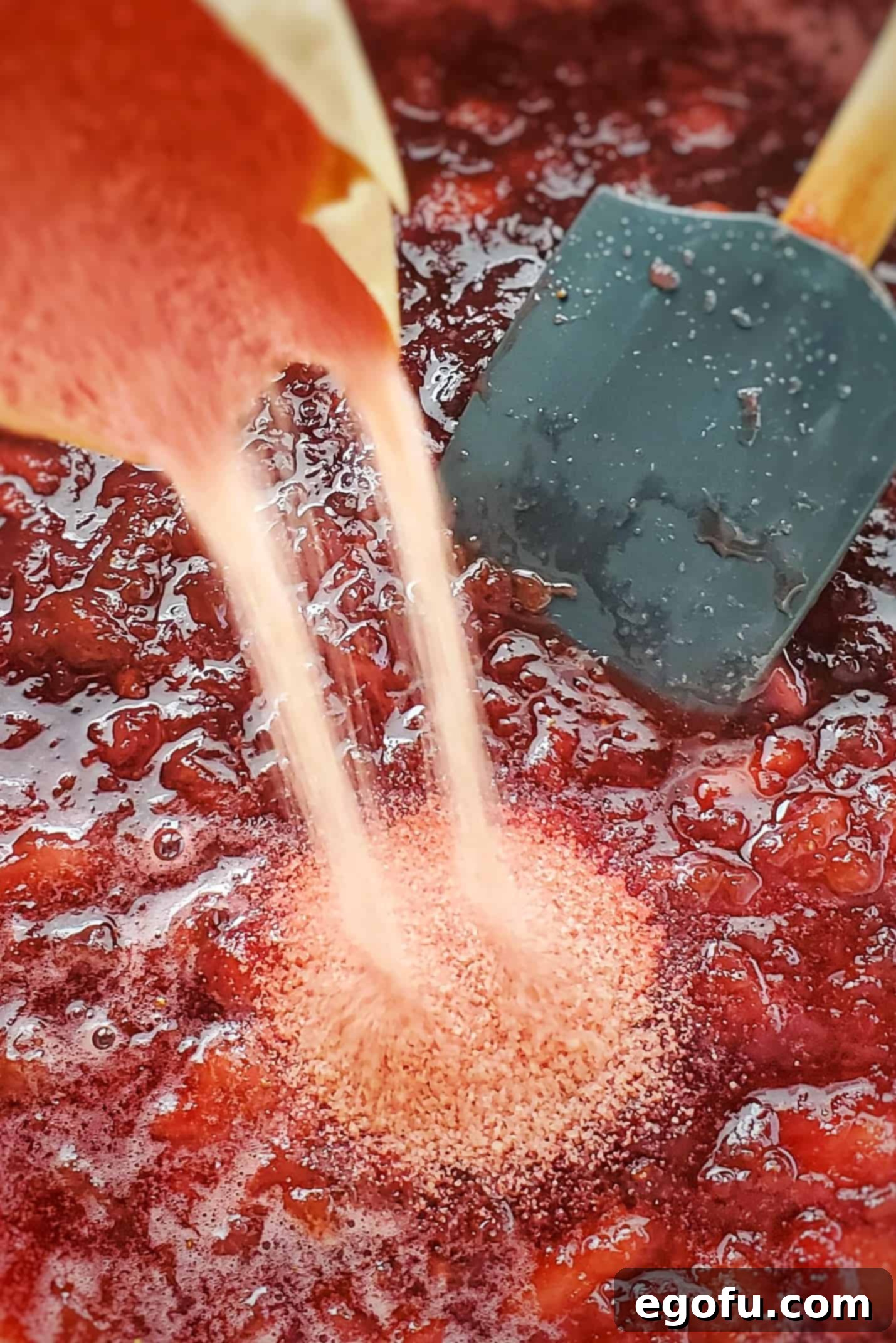 A hand pouring strawberry-flavored gelatin powder directly into the hot, just-removed-from-heat jam mixture in a saucepan.