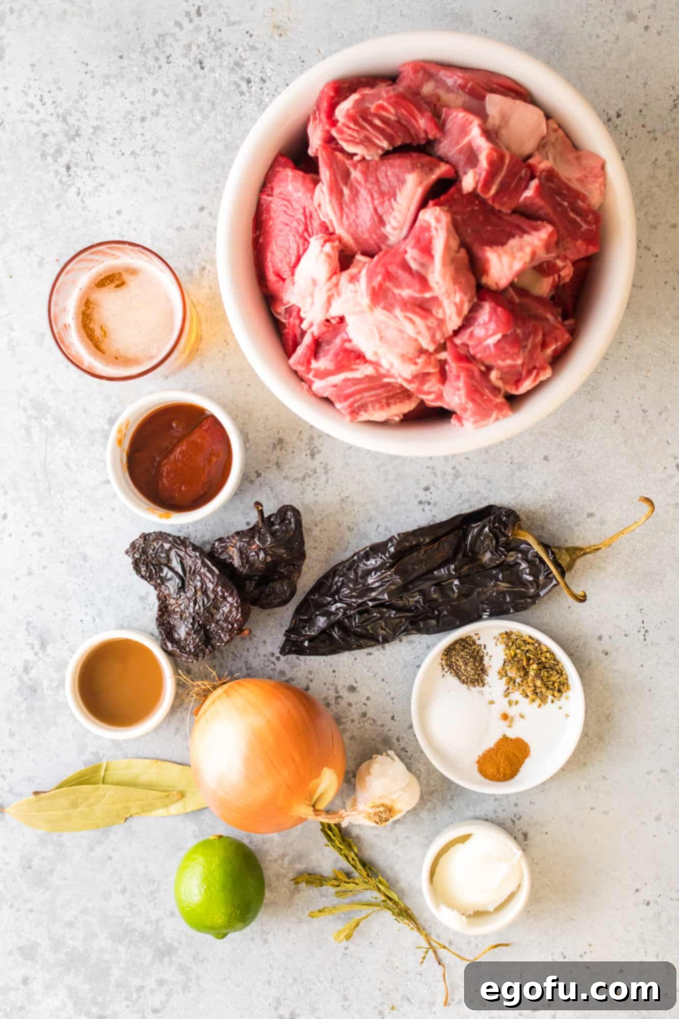 Various ingredients for Instant Pot Barbacoa laid out, including lard, olive oil, chuck roast, different types of chiles, lime, vinegar, spices, onion, garlic, and beer.