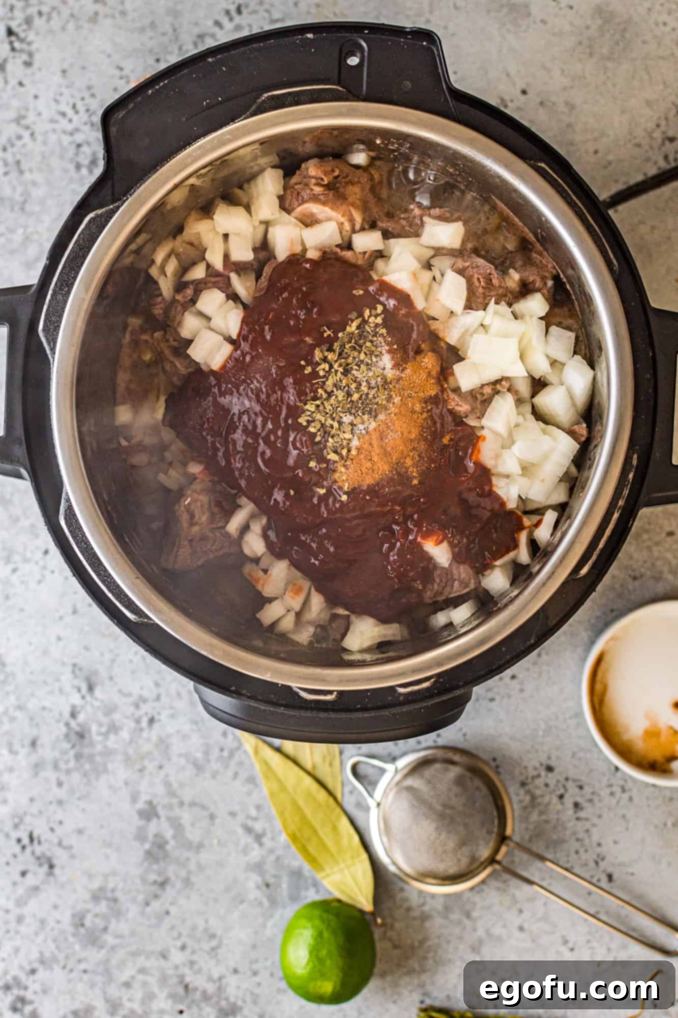Chili sauce, diced onion, bay leaves, epazote, oregano, and cinnamon added over seared meat in the Instant Pot, ready to be stirred.