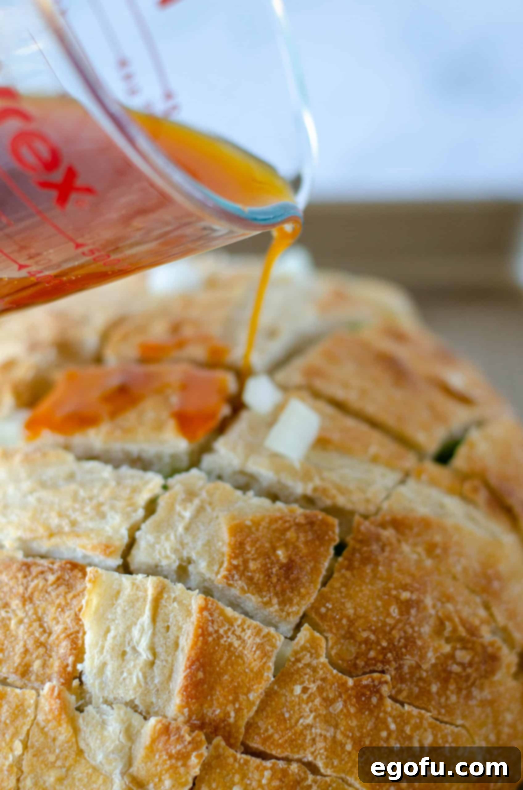 Measuring cup pouring the taco seasoning butter mixture over the cut and filled bread loaf on a baking sheet, ensuring even saturation.