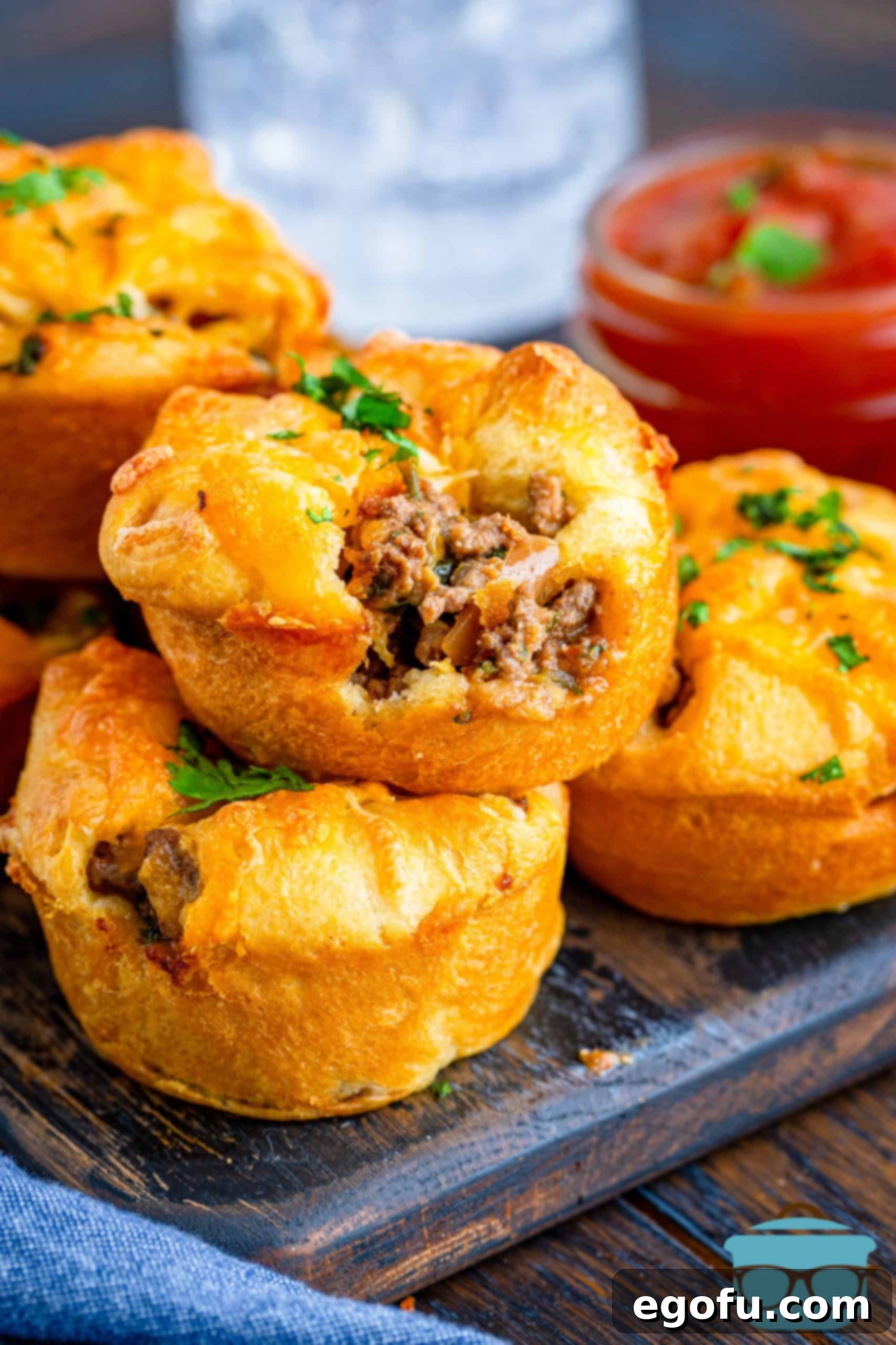 Stacked Crescent Taco Cups on wooden board with salsa in background, showcasing their golden crust and savory filling.