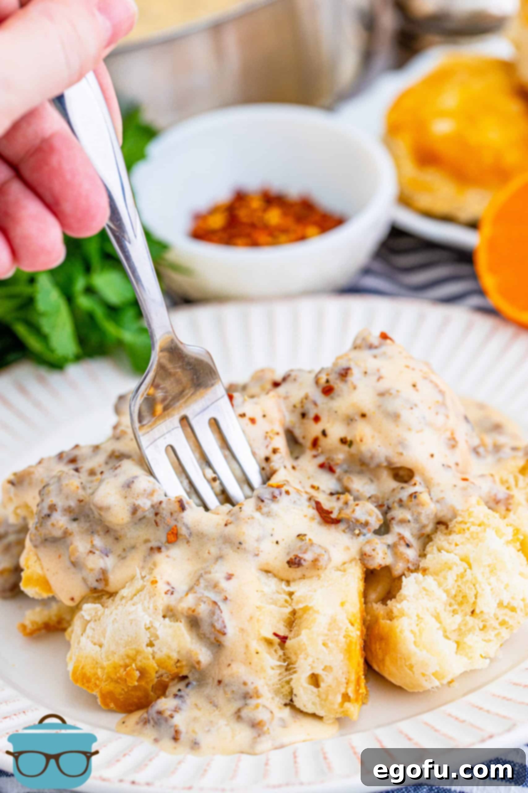 Homemade Sausage Gravy generously spooned over biscuits on a white plate, with a hand holding a fork ready to dig in.
