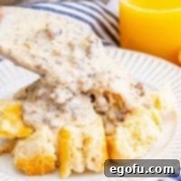 Close-up image of a wooden spoon scooping Homemade Sausage Gravy over biscuits on a white plate.