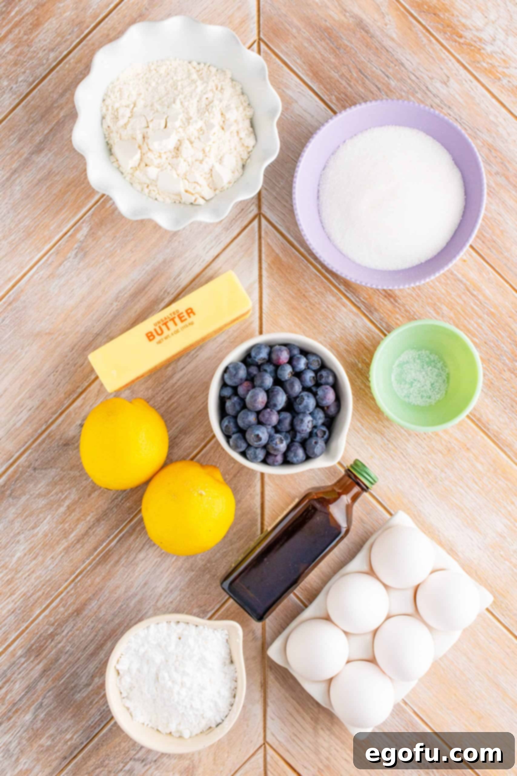 A flat lay of all the ingredients needed for Blueberry Lemon Bars: unsalted butter, granulated sugar, all-purpose flour, eggs, lemon zest, lemon juice, vanilla extract, fresh blueberries, and powdered sugar.