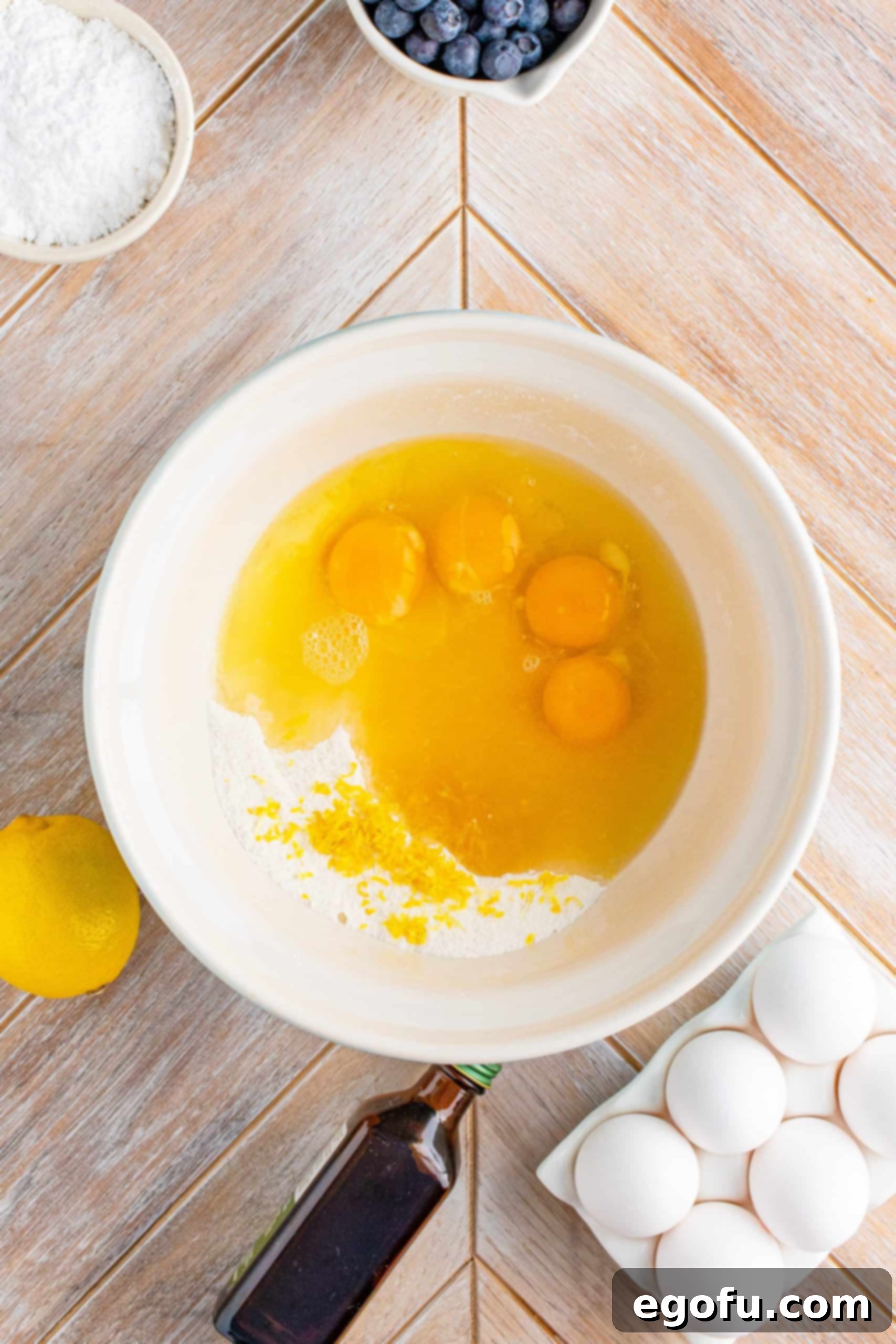 Eggs and lemon zest added to the flour and sugar mixture in a white bowl, ready to be whisked.
