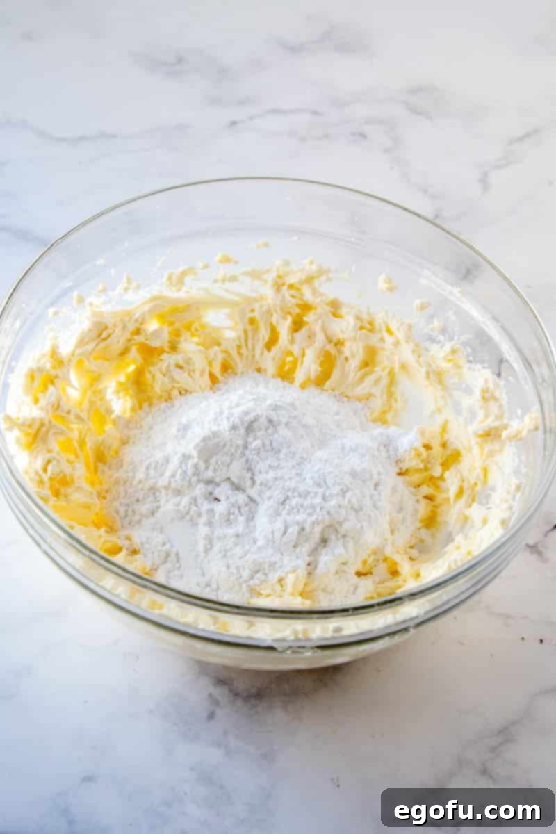Powdered sugar being added to the cream cheese and butter mixture in a clear bowl.