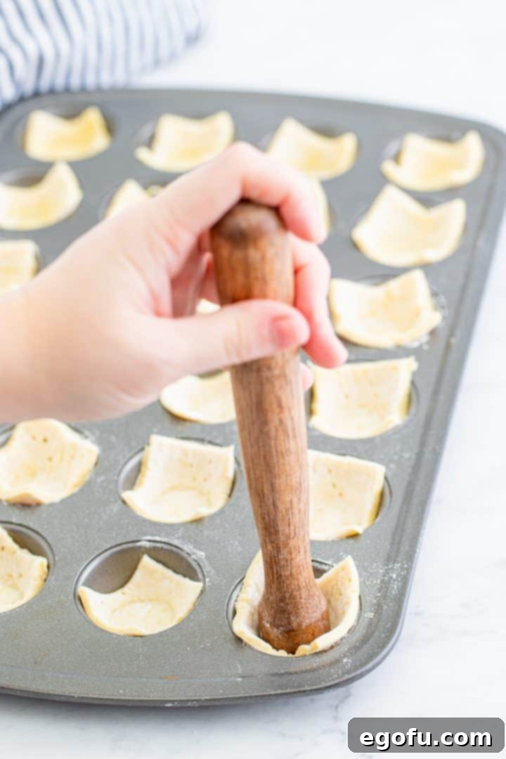 Cut puff pastry squares being meticulously pressed into a mini muffin tin's cavities.
