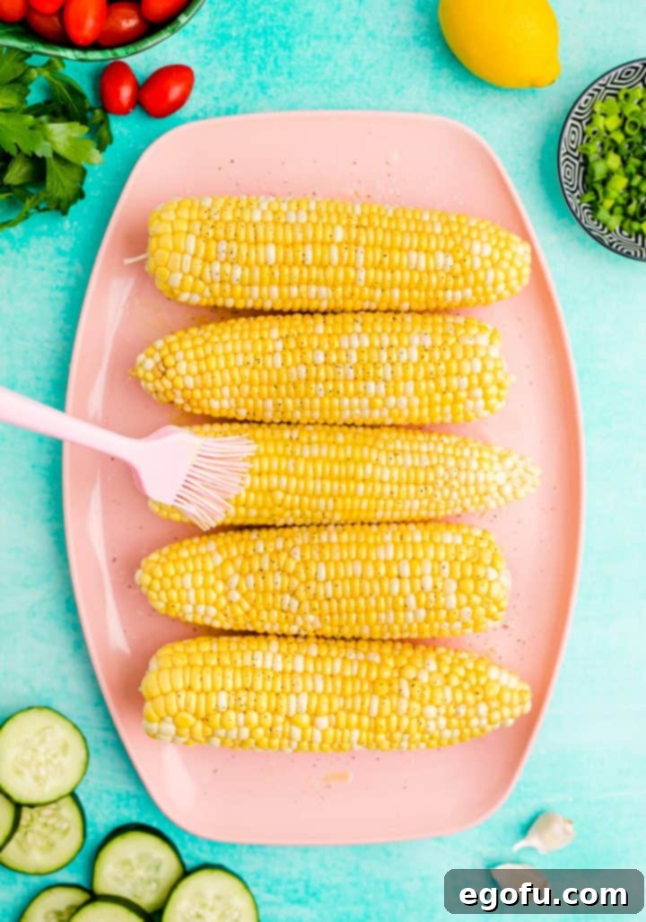 Corn cobs being brushed with olive oil and seasoned with salt and pepper