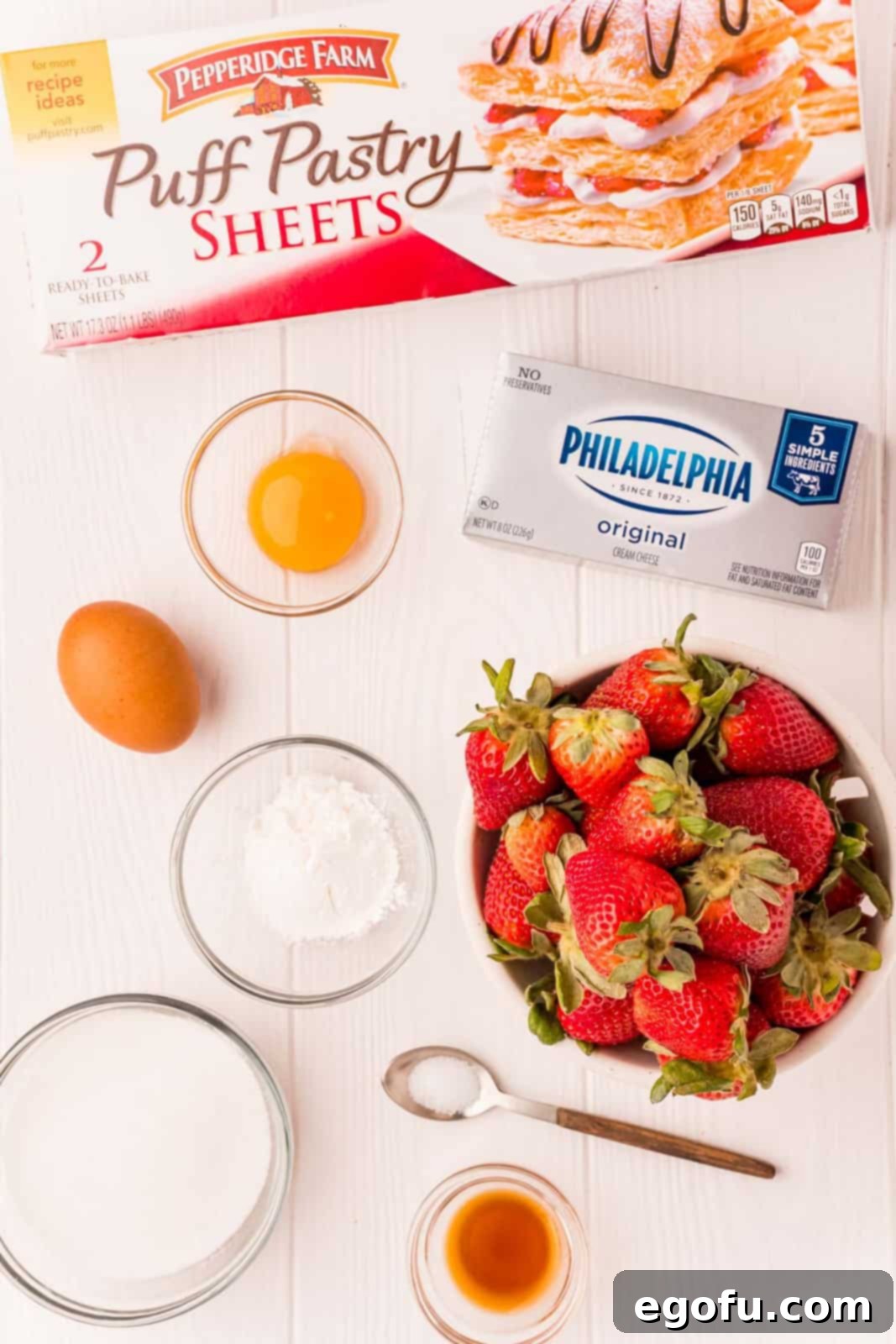 A flat lay image displaying all the ingredients needed for Strawberry Cheese Danishes: fresh strawberries, cornstarch, granulated sugar, puff pastry, cream cheese, egg yolk, vanilla extract, salt, a whole egg, water, and powdered sugar.