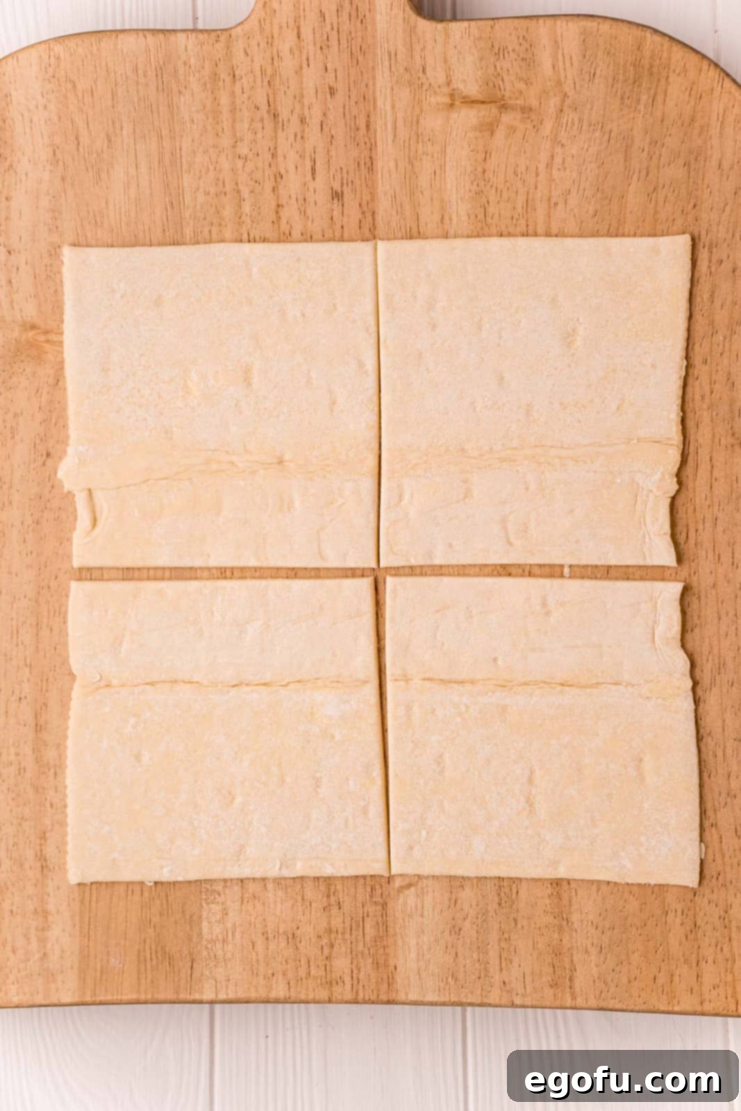 One sheet of puff pastry neatly cut into four perfect squares on a rustic wooden cutting board, ready for shaping.