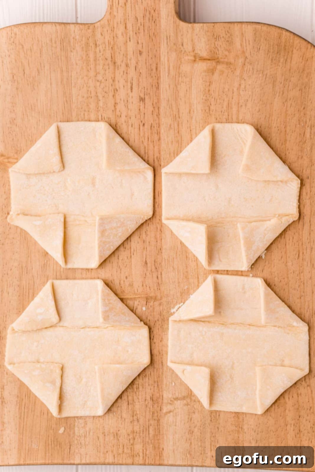 Puff pastry squares with their corners carefully folded inwards, creating the classic octagonal shape with a central well, resting on a wooden cutting board.