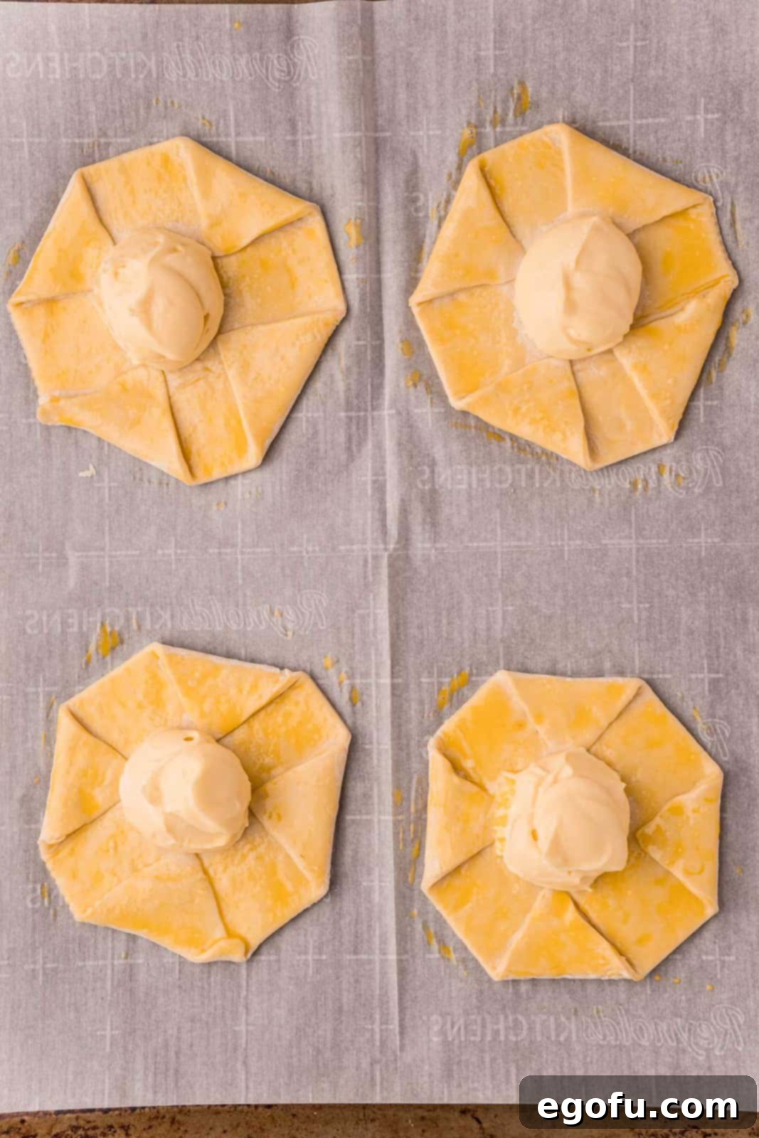 Unbaked danishes, meticulously topped with cream cheese mixture and brushed with a golden egg wash around the edges, resting on a parchment-lined baking sheet.