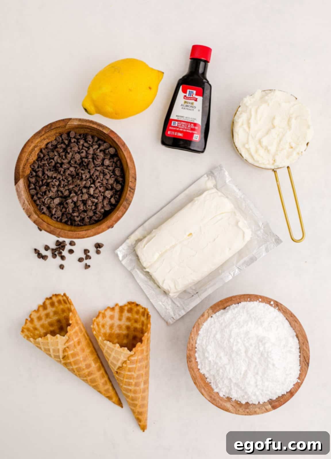 A vibrant flat lay image showcasing all the ingredients needed for Cannoli Dip: cream cheese, ricotta cheese, powdered sugar, almond extract, lemon zest, mini chocolate chips, and crisp waffle cones.