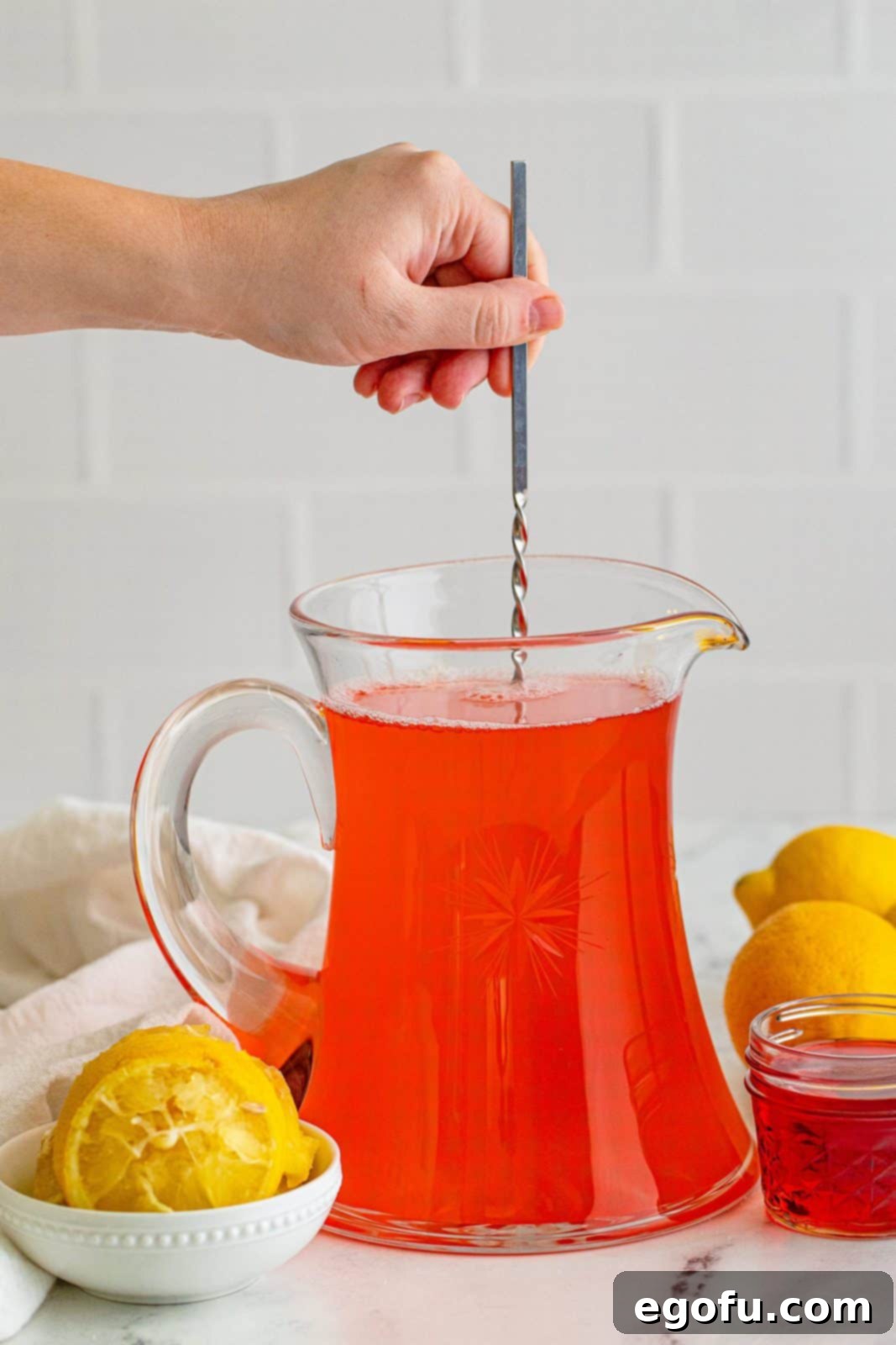 A hand stirring the Homemade Pink Lemonade in the pitcher, now fully combined and pink.