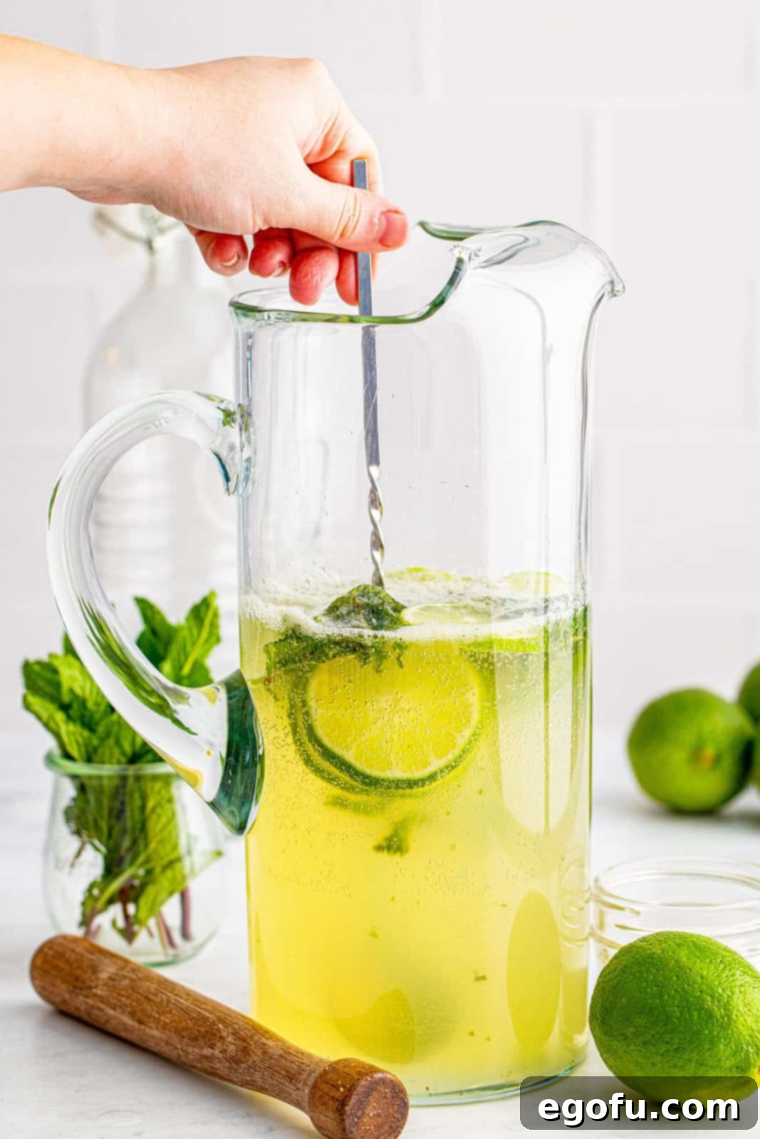 A long silver spoon gently stirring the fully mixed Mojito in a pitcher, ensuring all ingredients are combined without losing fizz.