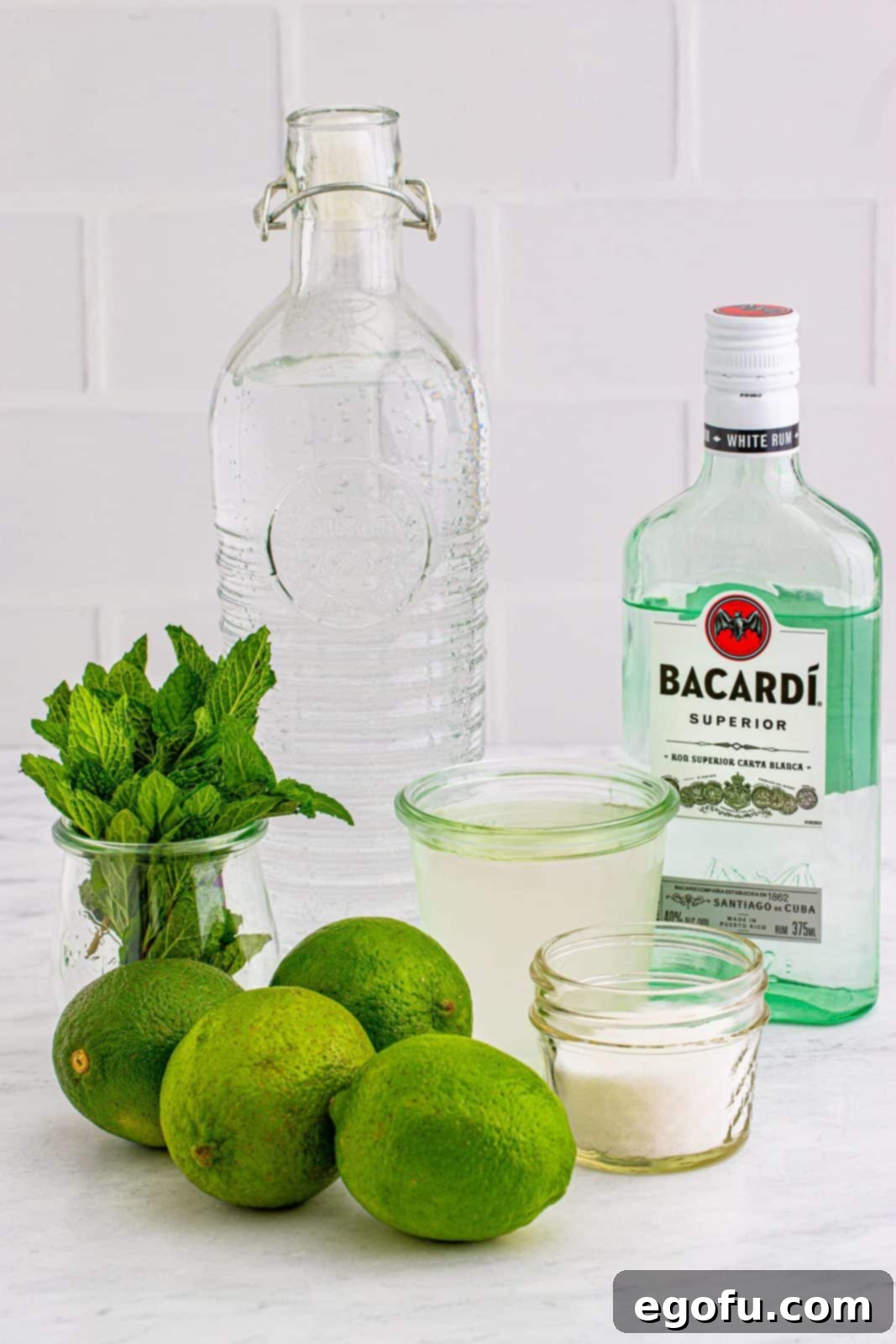 Arrangement of fresh ingredients for Summer Mojitos: whole limes, vibrant mint leaves, a bowl of granulated sugar, a jar of simple syrup, a bottle of white rum, and ice cubes.