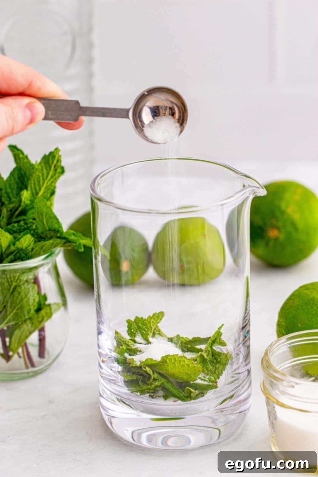 A glass pitcher on a countertop, ready for cocktail preparation, with fresh mint leaves and granulated sugar visible at the bottom.