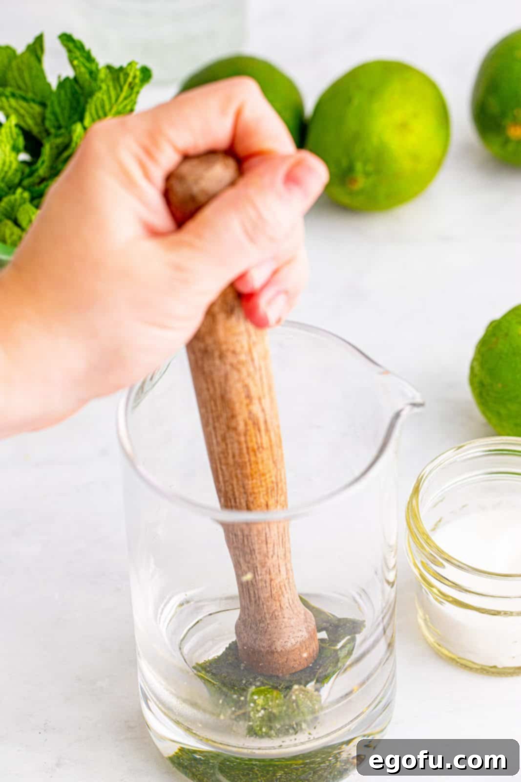 A hand using a muddler to gently crush fresh mint leaves and granulated sugar at the bottom of a clear glass cup, releasing their aromas.
