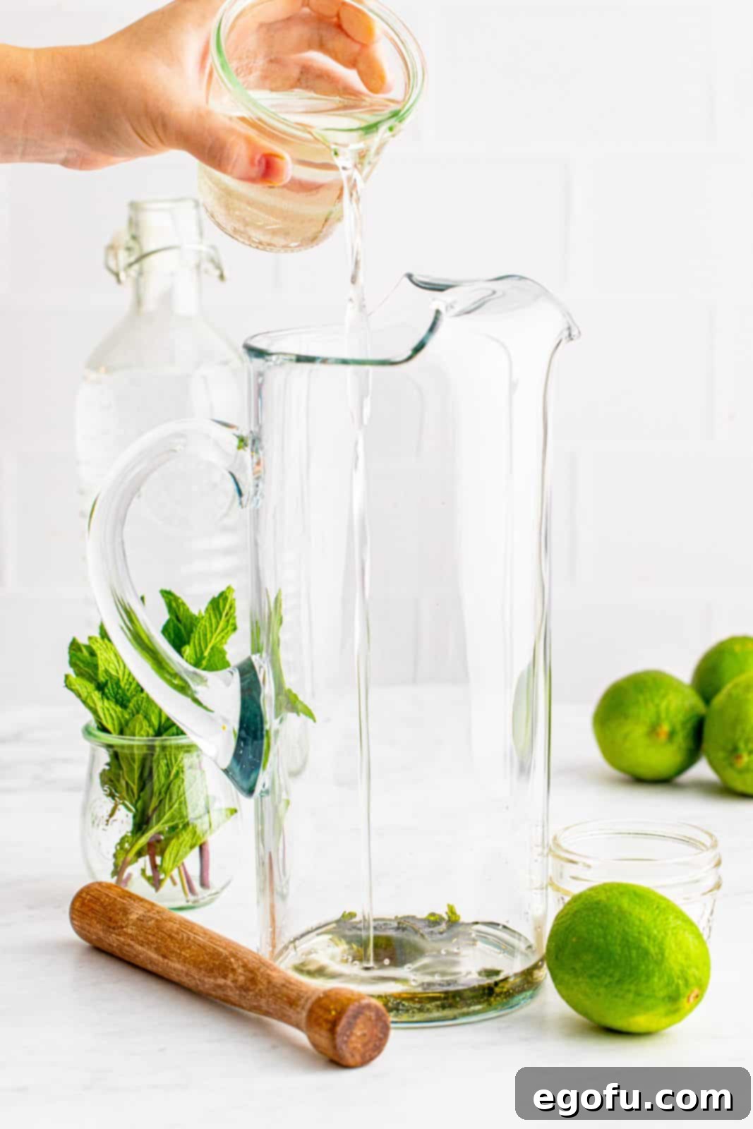 A stream of clear simple syrup being poured into a tall, transparent glass pitcher containing muddled mint.