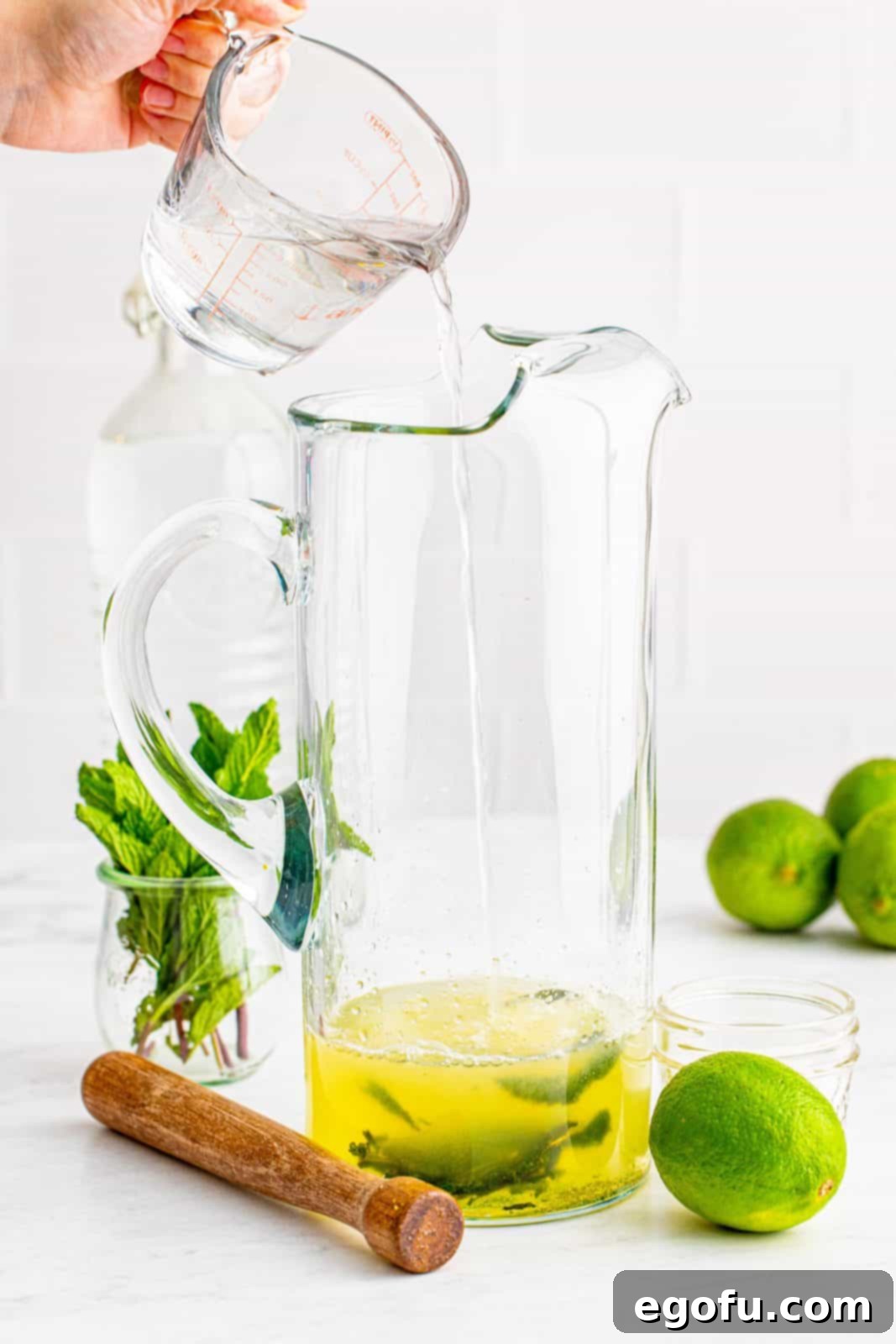 White rum being poured into a large glass pitcher, blending with the mint, sugar, simple syrup, and lime juice mixture.
