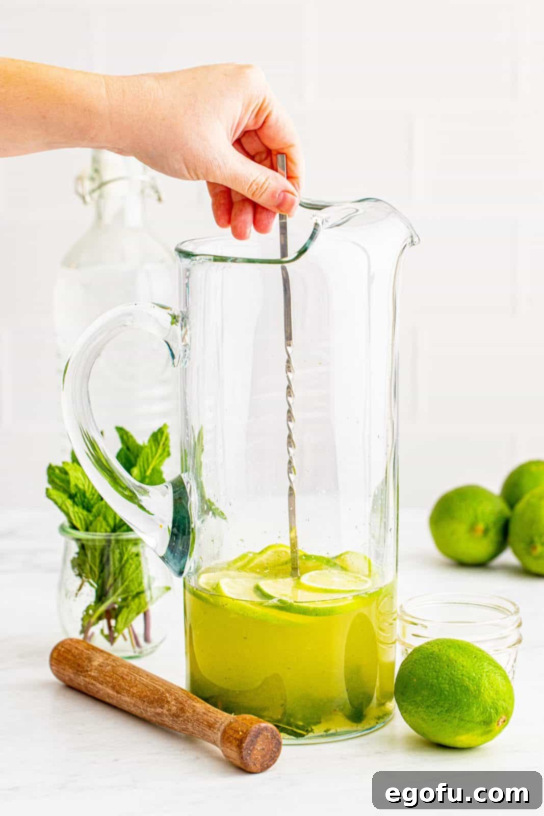 A long, silver drink stirrer being used to mix the Mojito base ingredients thoroughly in a tall glass pitcher.