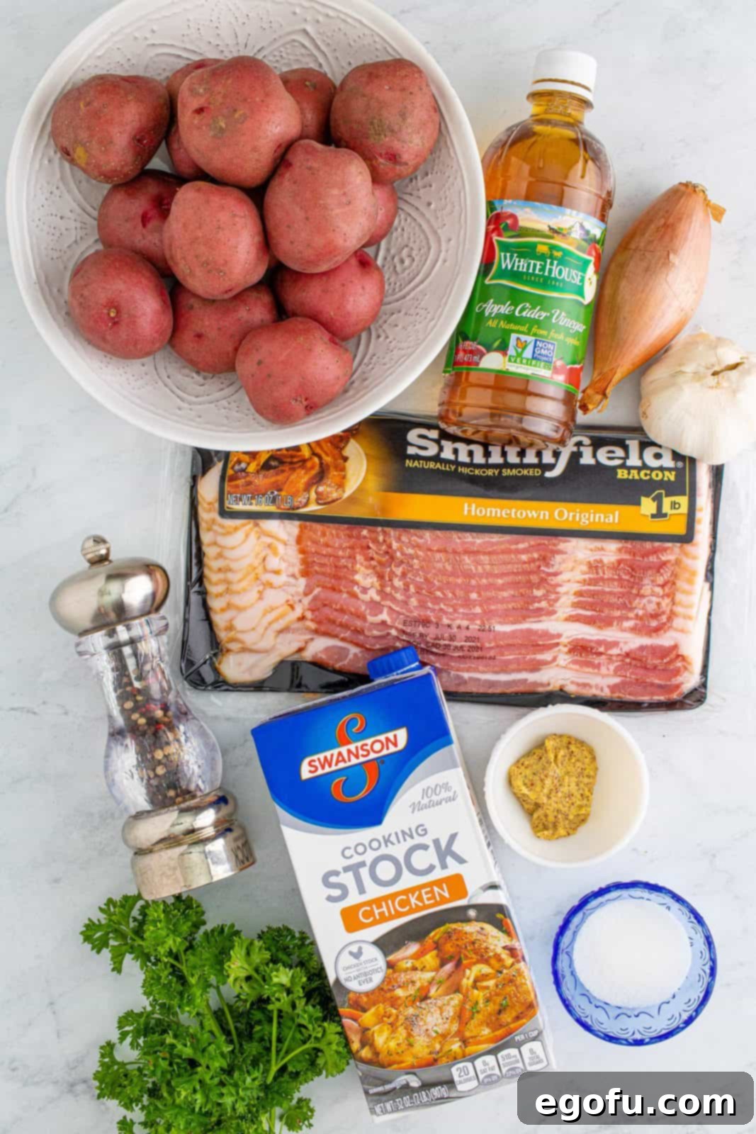 Hearty German Potato Salad 4 A collage of ingredients laid out: Red potatoes, raw bacon strips, a shallot, garlic cloves, bottles of apple cider vinegar and chicken stock, granulated sugar, whole grain mustard, a grinder of black pepper, and fresh parsley sprigs.