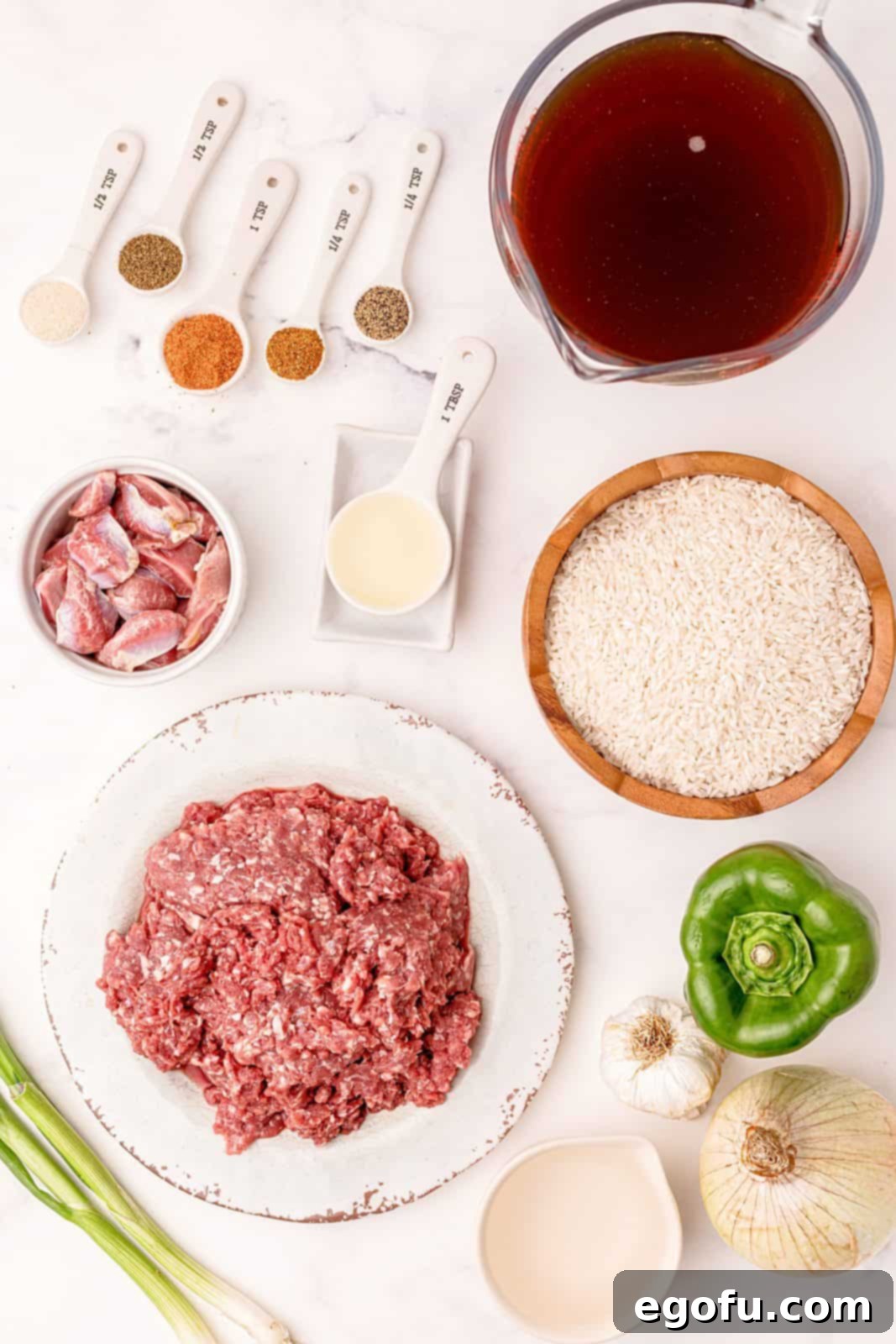 A beautifully arranged flat lay photo showcasing all the essential ingredients for making Dirty Rice, including long grain rice, beef broth, canola oil, lean ground beef, chicken gizzards, green bell pepper, sweet onion, garlic, celery seed, Creole seasoning, onion powder, cayenne pepper, ground black pepper, water, and fresh green onions.