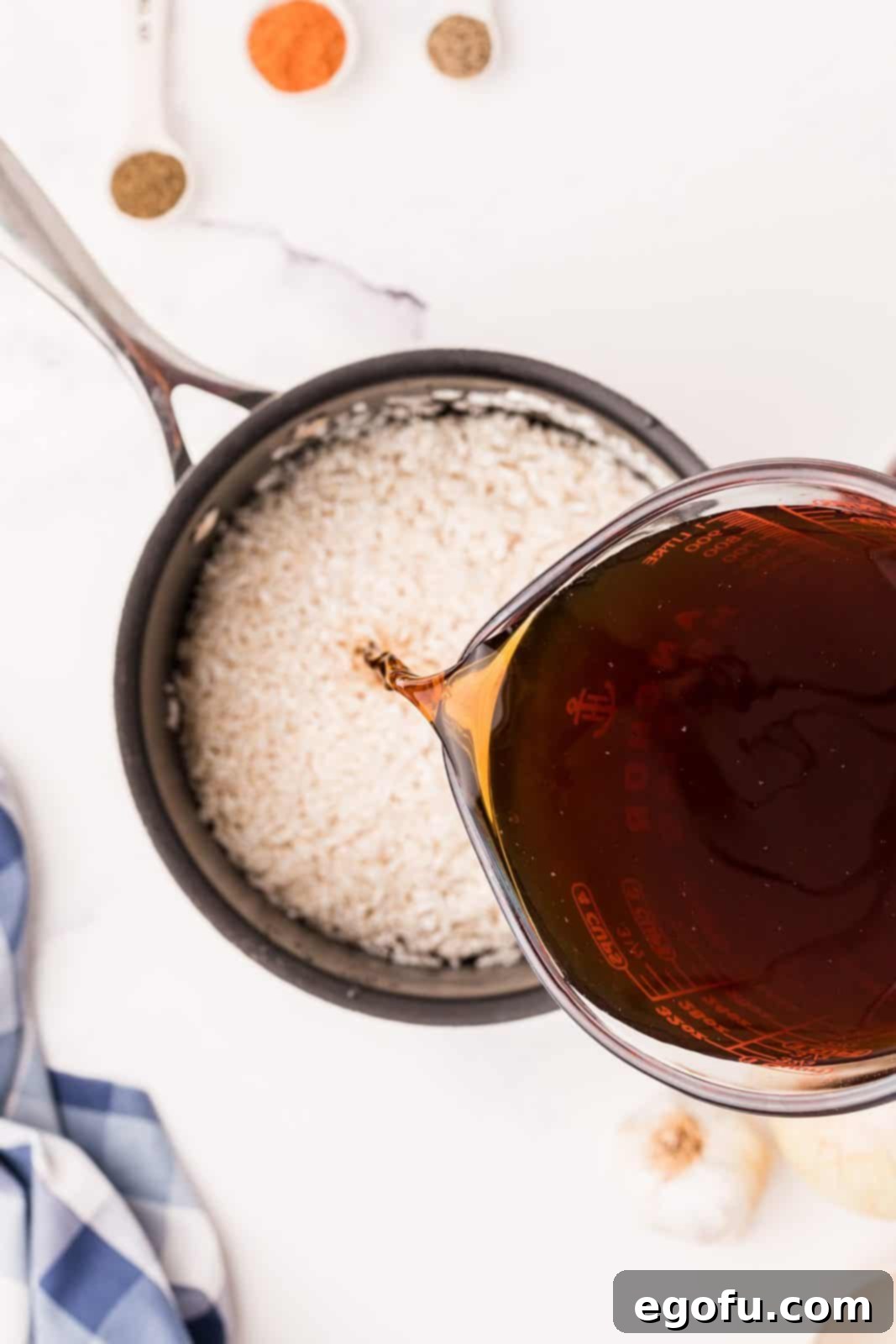 Close-up shot of rich beef broth being poured over dry long grain rice in a saucepan, initiating the cooking process for the Dirty Rice.