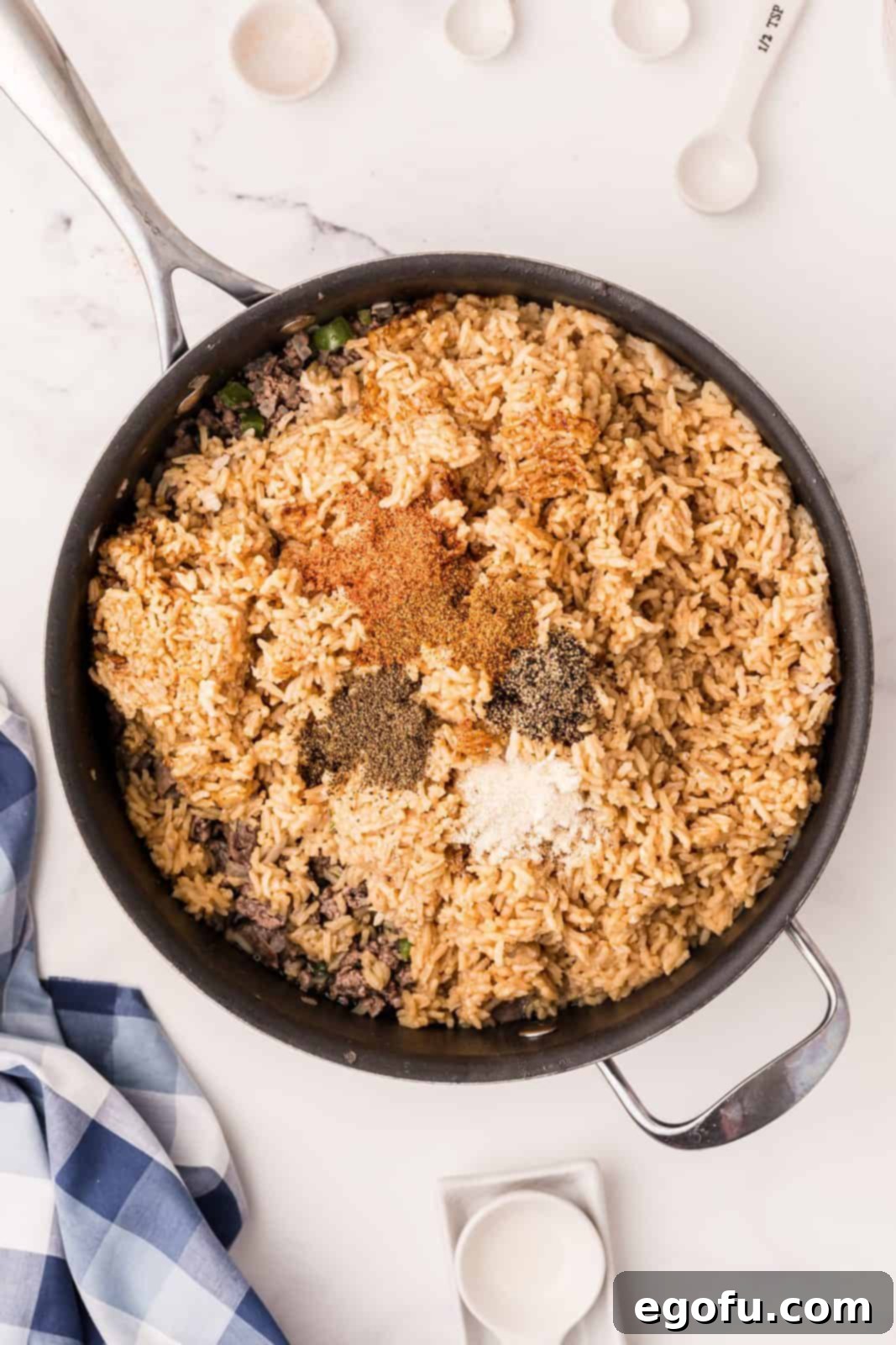 A mix of cooked meat and vegetables in a skillet, now combined with a vibrant array of spices including celery seed, Creole seasoning, onion powder, cayenne pepper, black pepper, and water, being stirred to incorporate flavors evenly.