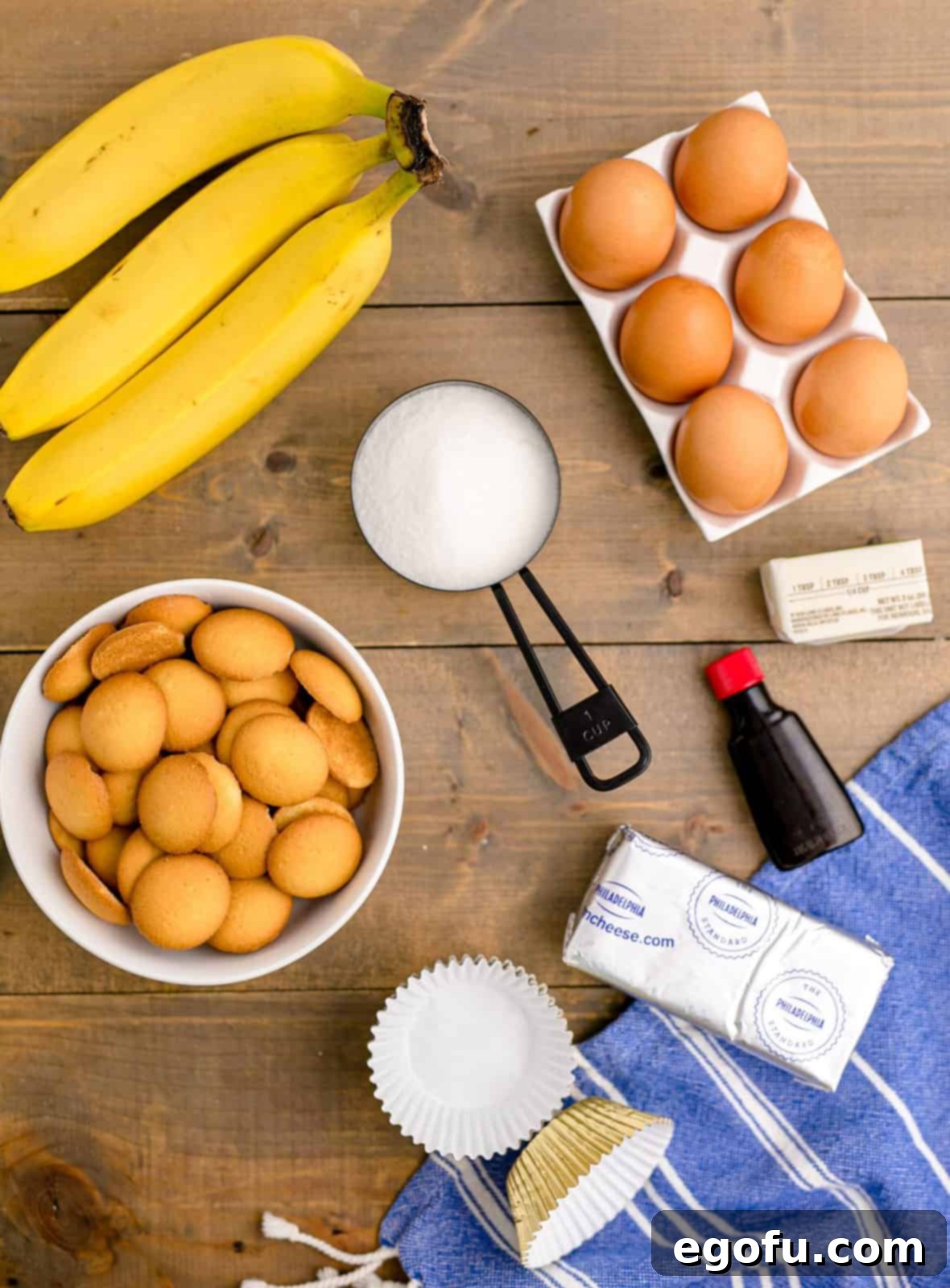 Ingredients needed for Mini Banana Pudding Cheesecakes: vanilla wafer cookies, salted butter, cream cheese, granulated sugar, eggs, vanilla, bananas, and whipped cream, neatly arranged.