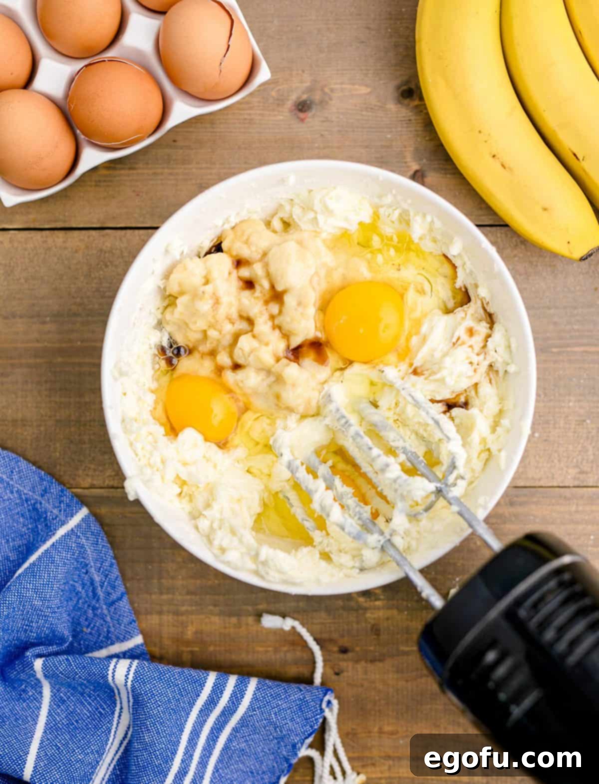 Eggs, vanilla, and mashed banana added to the cream cheese mixture in a white bowl, ready for final mixing.