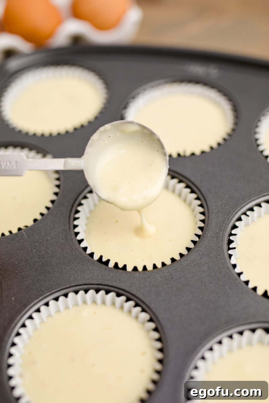 Cheesecake filling being spooned into paper liners in a muffin tin, demonstrating the filling process.